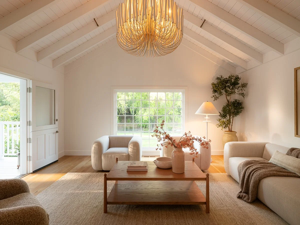 Bright living room with large window, beige armchair, white sofa with blanket, wooden coffee table with vases and books, floor lamp, potted plant, chandelier, door to outdoor balcony, neutral decor, hardwood floors.