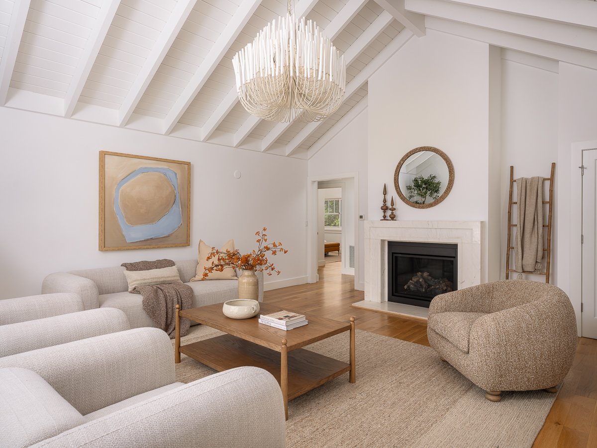 Living room with white walls, a vaulted ceiling with white beams, a chandelier, a fireplace with a mirror above it, beige and gray furniture, a wooden coffee table, and decorative items including a vase with orange flowers.