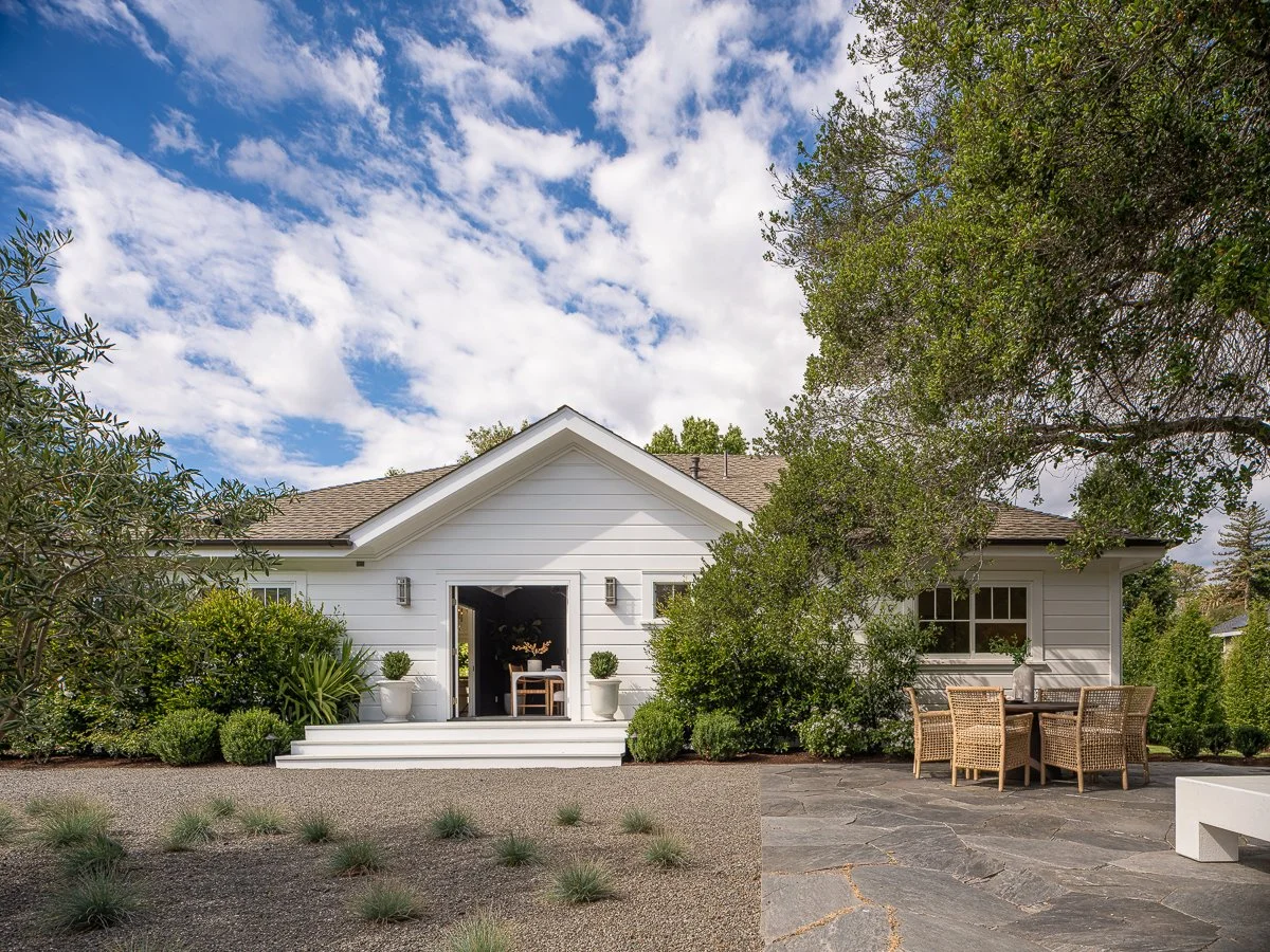 White house with a front porch, outdoor chairs, green shrubs, trees, and a partly cloudy sky.