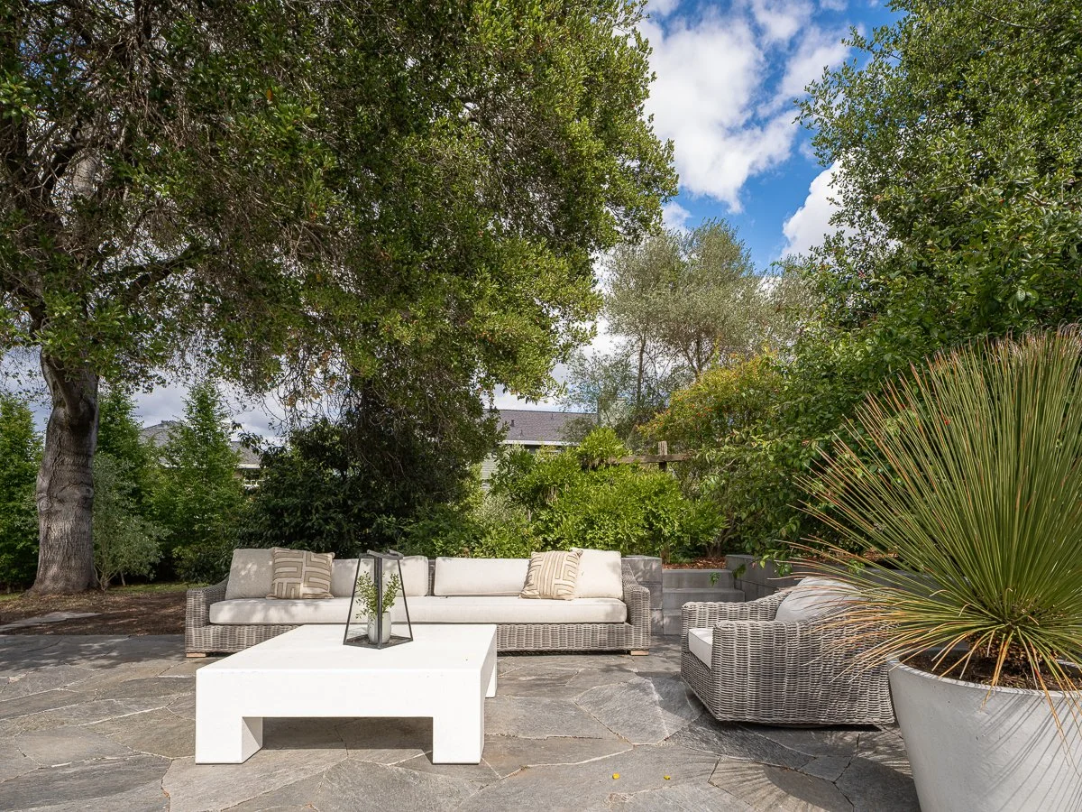 Outdoor patio with wicker sofa, armchair, white coffee table, potted plants, and trees under a partly cloudy sky.