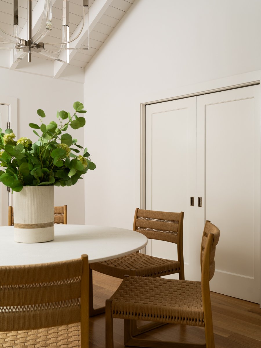 A dining room with a round white table, four wooden chairs, a large potted plant with green leaves, white walls, a white ceiling with exposed beams, and sliding closet doors.
