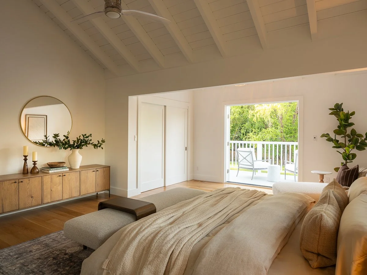 Living room with a tropical decor, beige sofa, wooden sideboard with candles and a mirror, patio doors leading to a deck with outdoor seating, trees outside.