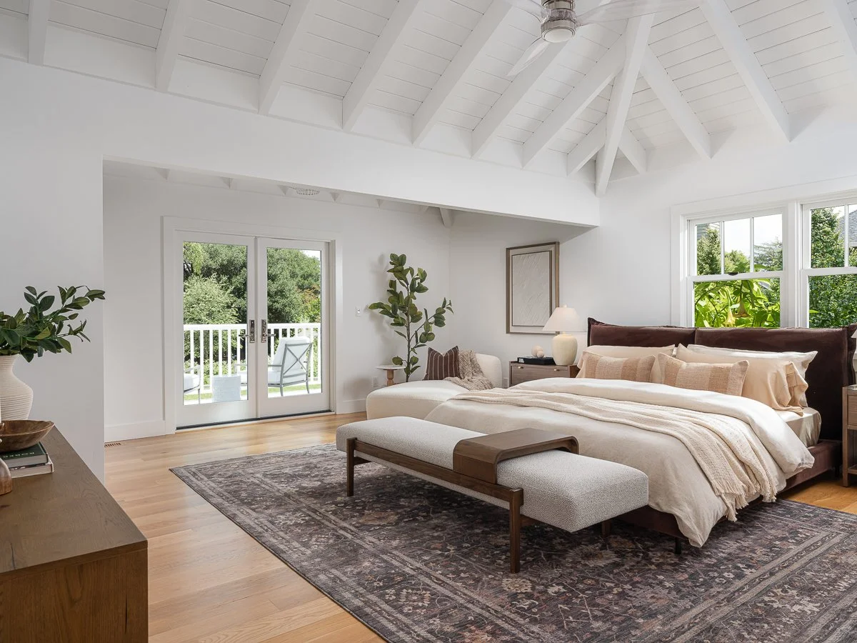 A bedroom with white walls, a vaulted ceiling, large windows, and a balcony door, furnished with a bed, bench, potted plants, and a rug.