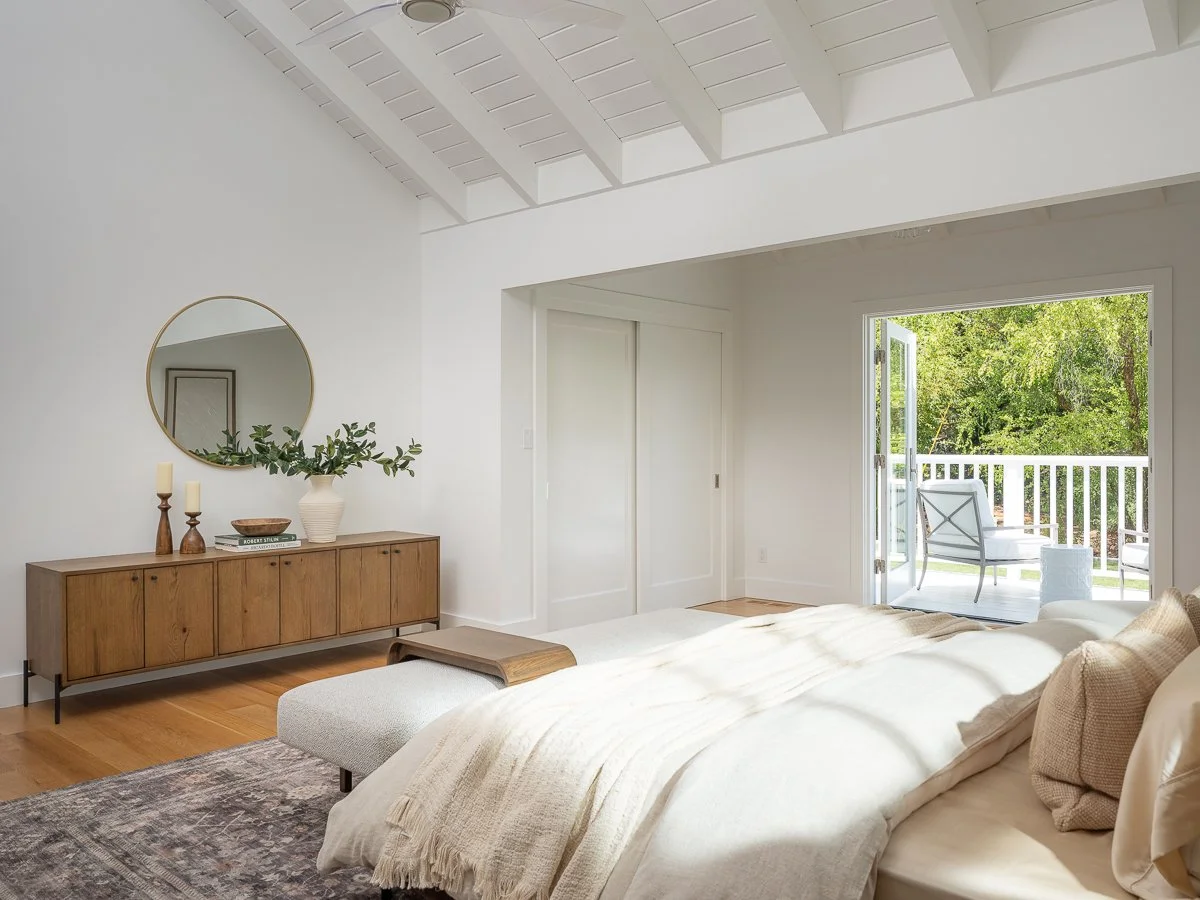Bright, minimalist bedroom with white walls and ceiling, a wooden sideboard with plants and candles, a large mirror, a bed with beige linens, and a sliding glass door leading to a balcony with outdoor chairs and surrounded by greenery.