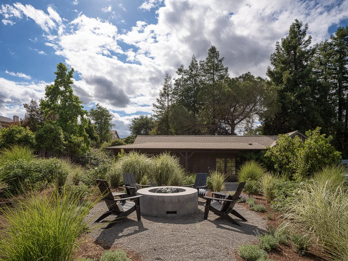 A backyard scene with a firepit surrounded by four black Adirondack chairs, lush greenery, and tall trees under a partly cloudy sky.