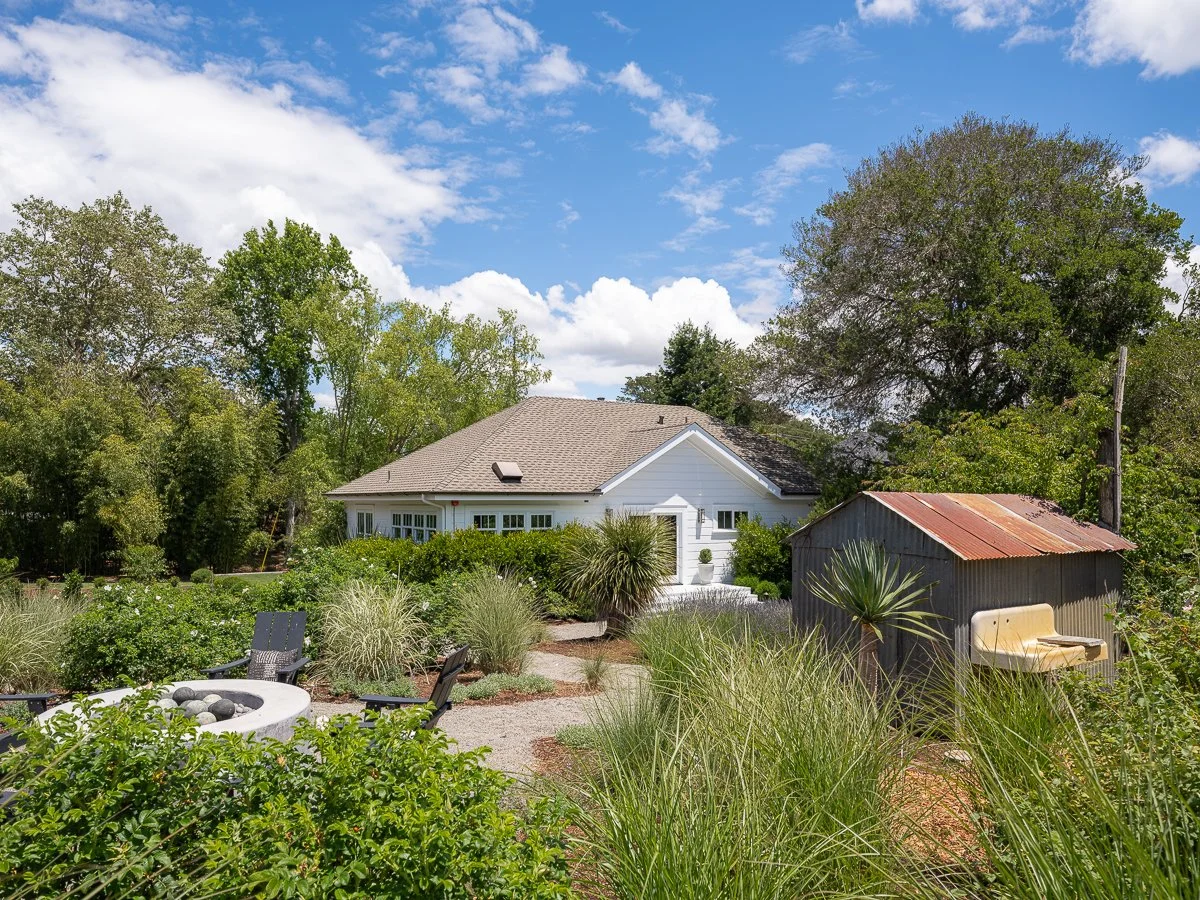 A backyard garden with a white house, a small shed with a rusted metal roof, surrounding green plants, trees, a fire pit with chairs, and a blue sky with scattered clouds.