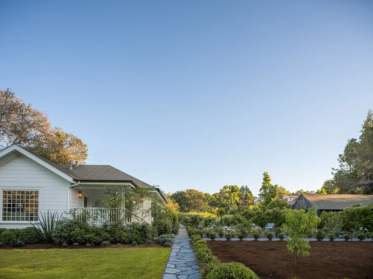 Residential house with landscaped garden, stone pathway, and neighboring homes under a blue sky.