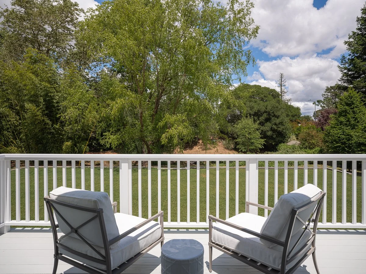 View of a backyard with two chairs on a white deck, greenery and trees in the background, blue sky with clouds.
