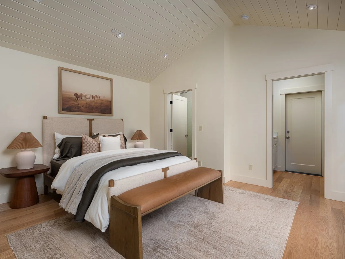 A bedroom with a bed and a bench, two nightstands with lamps, a framed picture of horses, and a doorway leading to a bathroom.