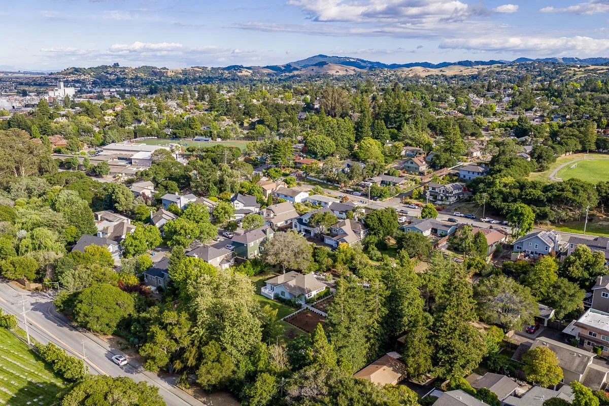 Aerial view of a residential neighborhood with houses, trees, and streets, with hills in the distance under a partly cloudy sky.