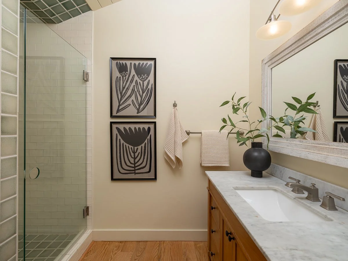 A bathroom with a glass shower, wall art, a towel rack with towels, a vase with green leaves, a mirror, and a wooden vanity with a marble countertop.