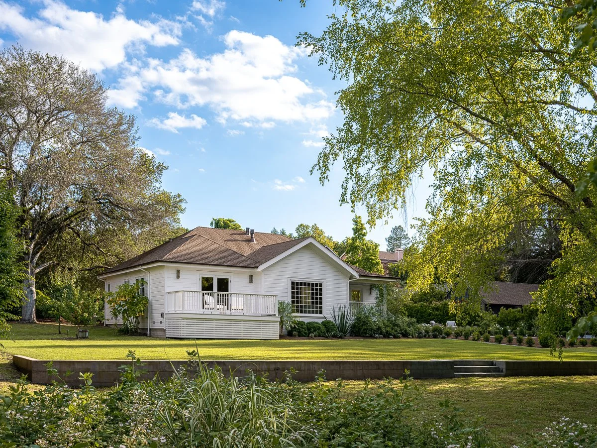 A white house with a brown roof surrounded by green trees and a well-maintained lawn under a partly cloudy sky.