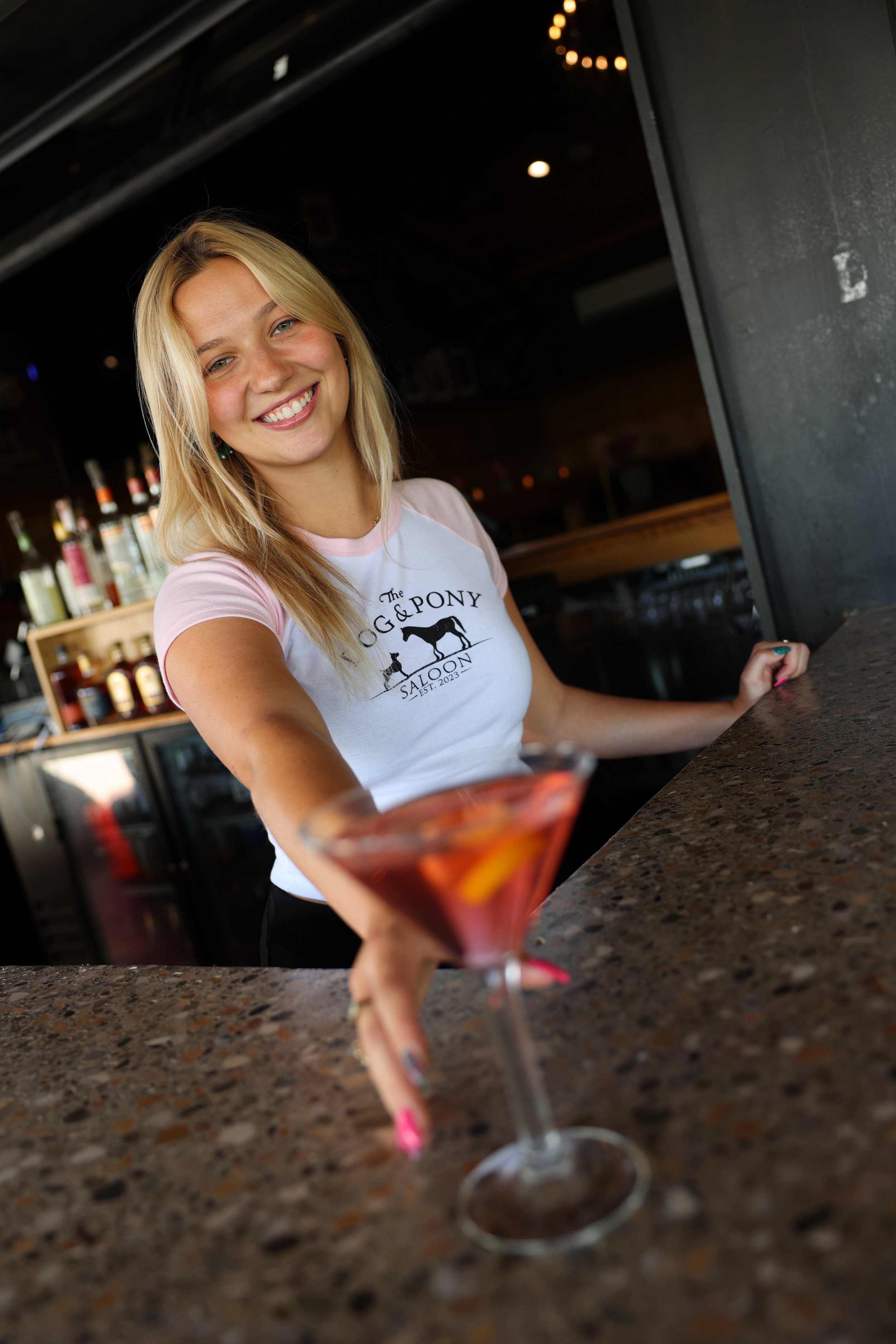A smiling blonde woman in a light pink T-shirt is at a bar, reaching out towards a pink cocktail in the foreground, with liquor bottles behind her.