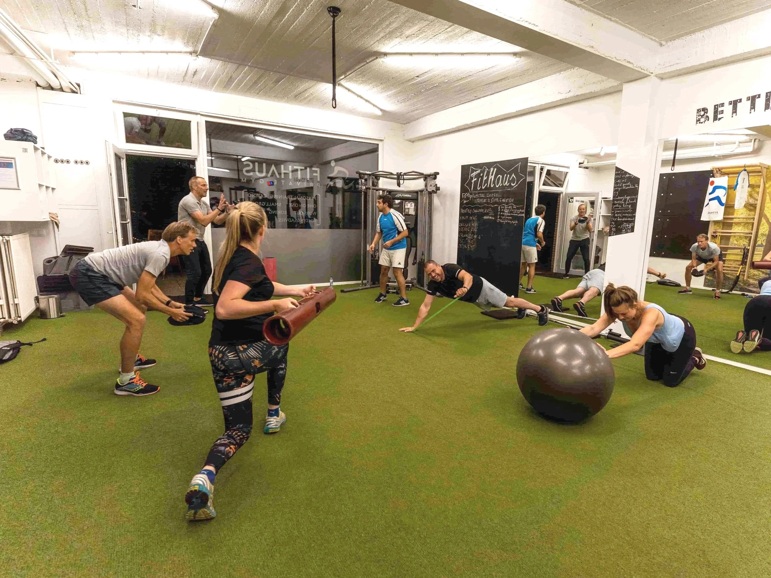 Groep mensen die in een fitnessruimte trainen, bezig met verschillende oefeningen zoals stretchen en gebruik van fitnessapparatuur, binnen een sportschool met groene vloer en lichte muren.