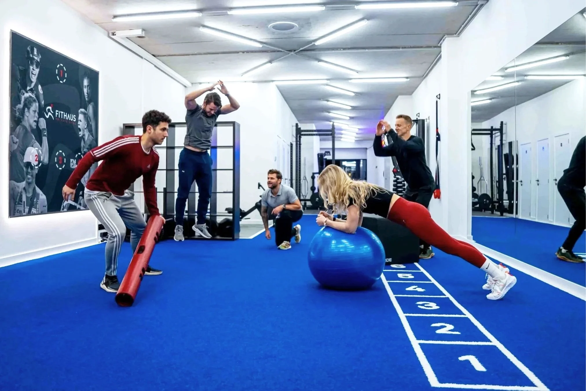Mensen trainen in een sportschool met fitnessequipment, zoals een grote blauwe fitnessbal en foam rollers, op een blauwe mat met nummering, terwijl een vrouw ondersteuning geeft tijdens een plankoefening.