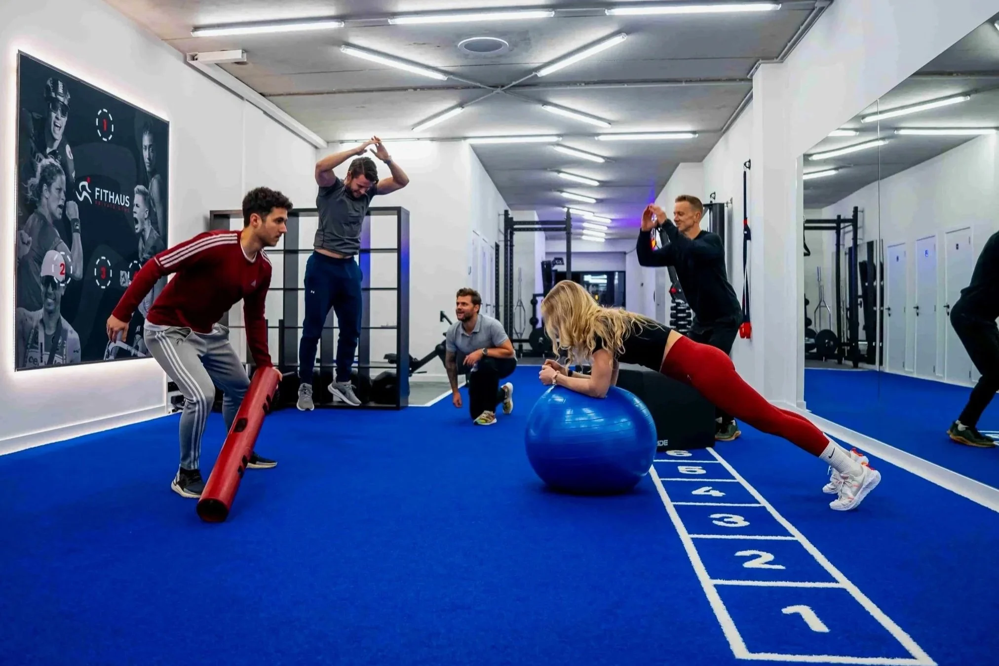 Mensen trainen in een sportzaal met blauwe matten, mede bezig met various fitness oefeningen, waaronder plank en strekoefeningen, onder begeleiding van een trainer.
