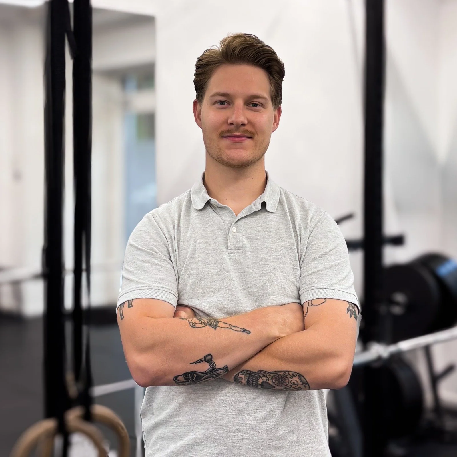 Een jonge man met getatoeëerde armen, gekleed in een lichtgrijze polo, staat met armen crossed in een fitnessruimte met cardio-apparatuur op de achtergrond.