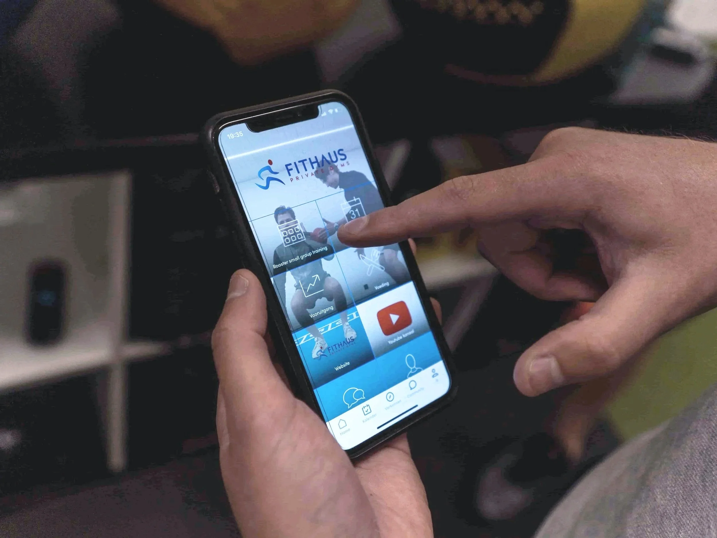 Een persoon houdt een smartphone met een fitness app op het scherm, met de tekst 'Fithaus' en afbeeldingen van mensen die trainen.