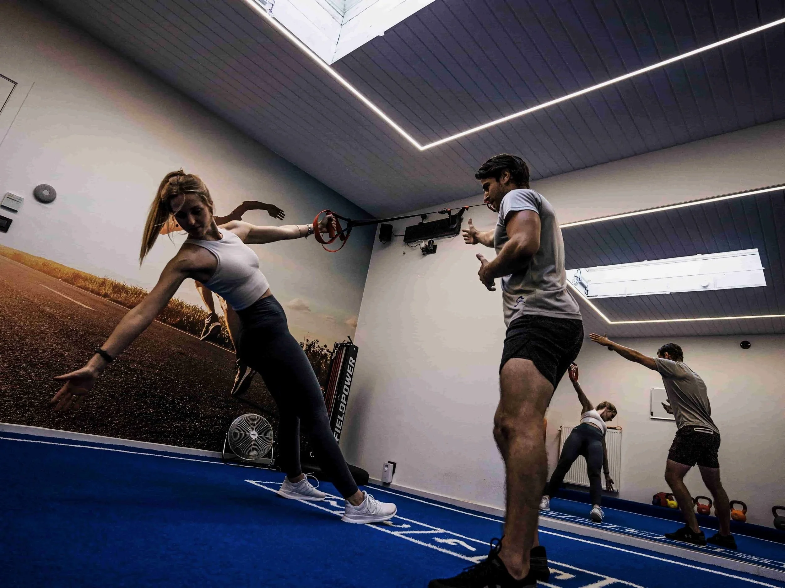 Binnen in een sportschool, mensen doen stretching oefeningen op een blauwe mat, met een muur waarop een afbeelding van een weg en een veld te zien is. Mensen zijn in een groep bezig met fitnessoefeningen.