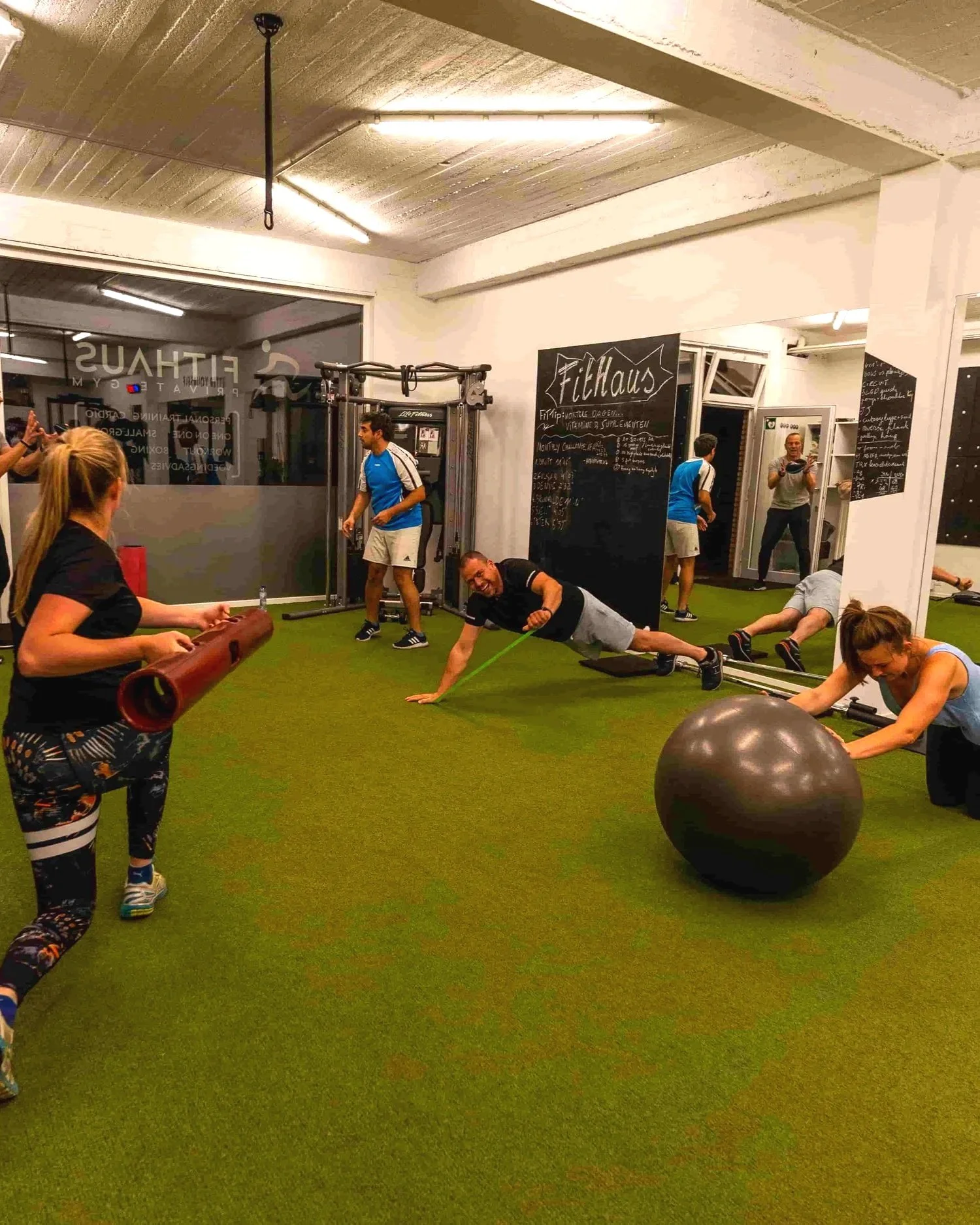 Binnen in een sportschool met mensen die oefeningen doen en trainen, inclusief een persoon die een plankhouding uitvoert, een grote balancebal en een verticaal zwart bord met het woord 'FitHaus' geschreven.