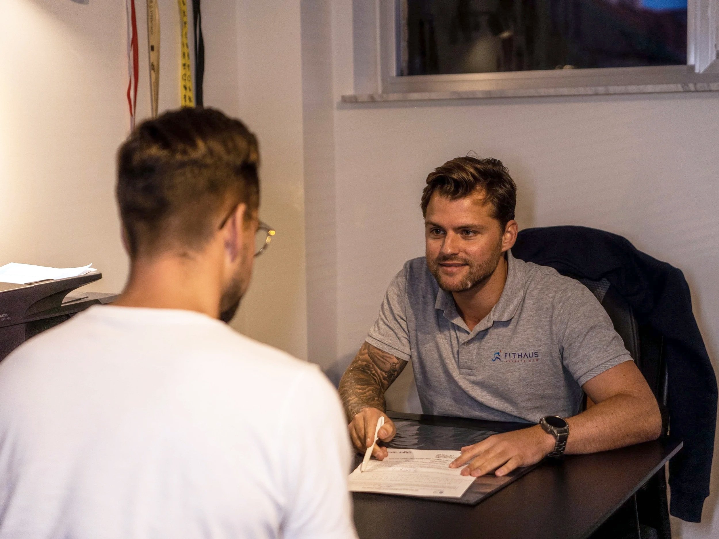 Twee mannen zitten aan een tafel, de man aan de rechterkant heeft een grijze polo met het logo 'FITHAUS' en een horloge en is bezig met het invullen van papieren, de andere man is van achter te zien.
