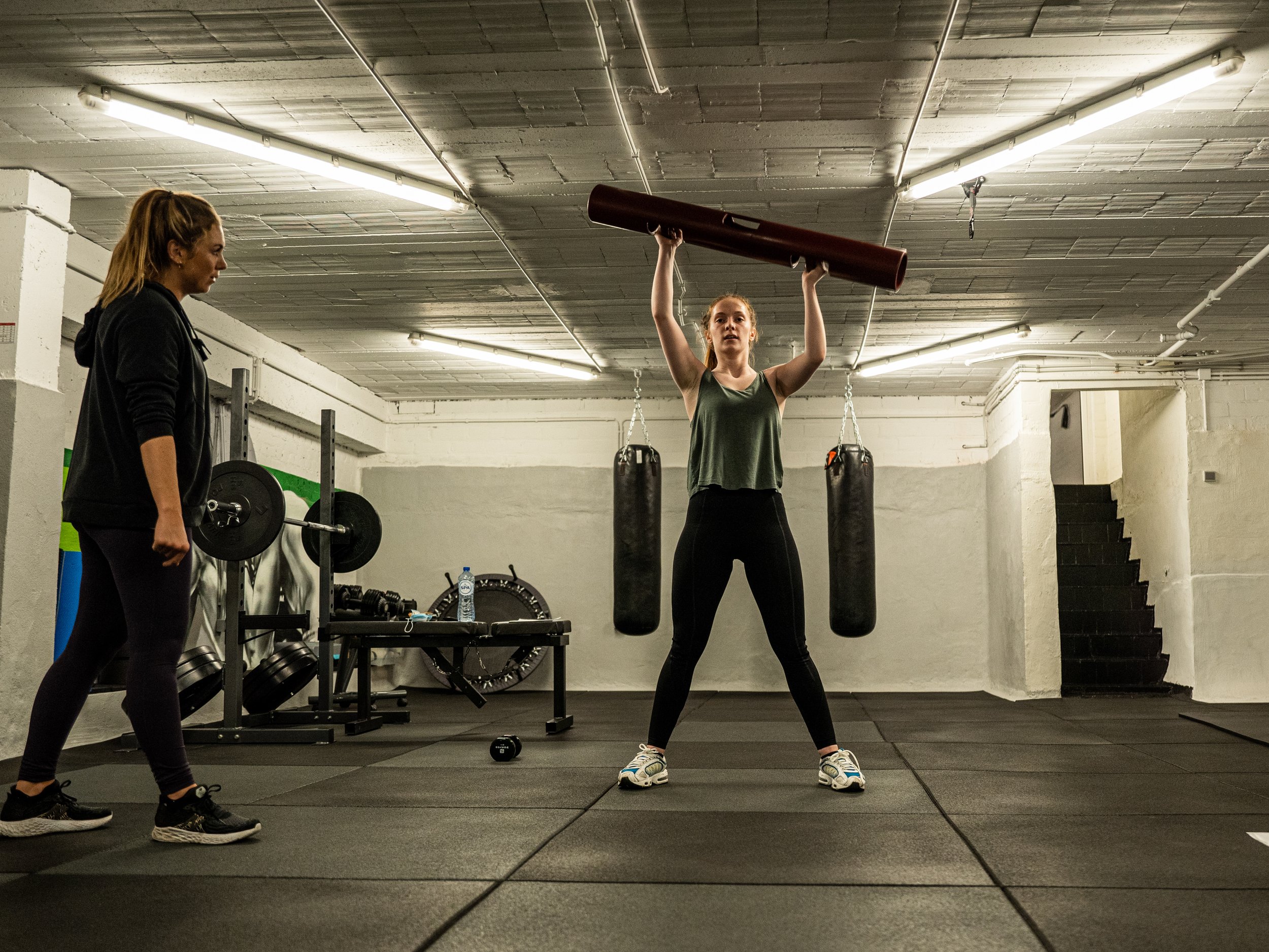 Een vrouw in sportkleding tilt een houten buis boven haar hoofd in een fitnessruimte, terwijl een andere vrouw haar observeert.