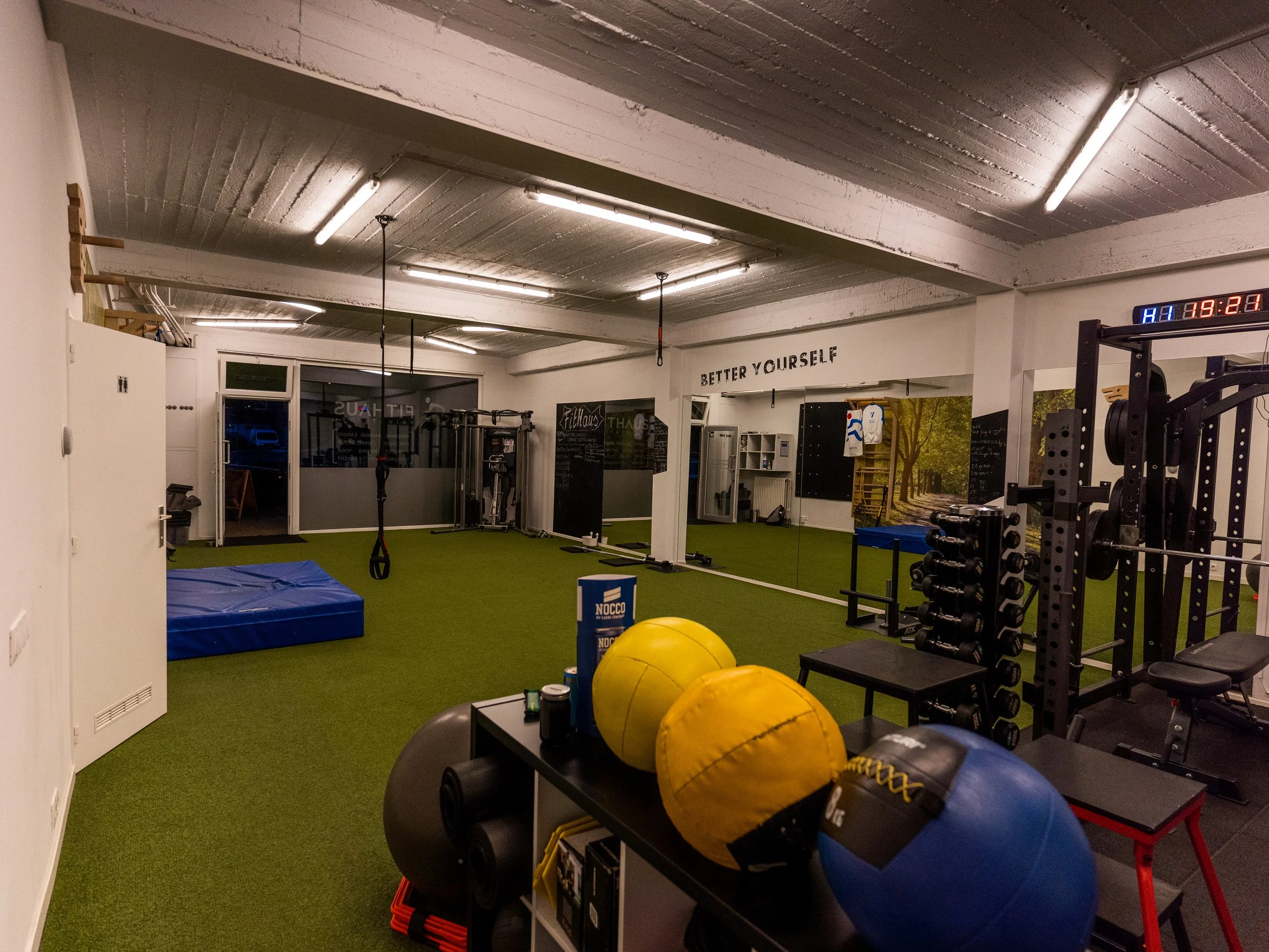 Binnenkant van een sportschool met fitnessapparatuur, medische ballen, posters en een digitale klok aan het plafond.