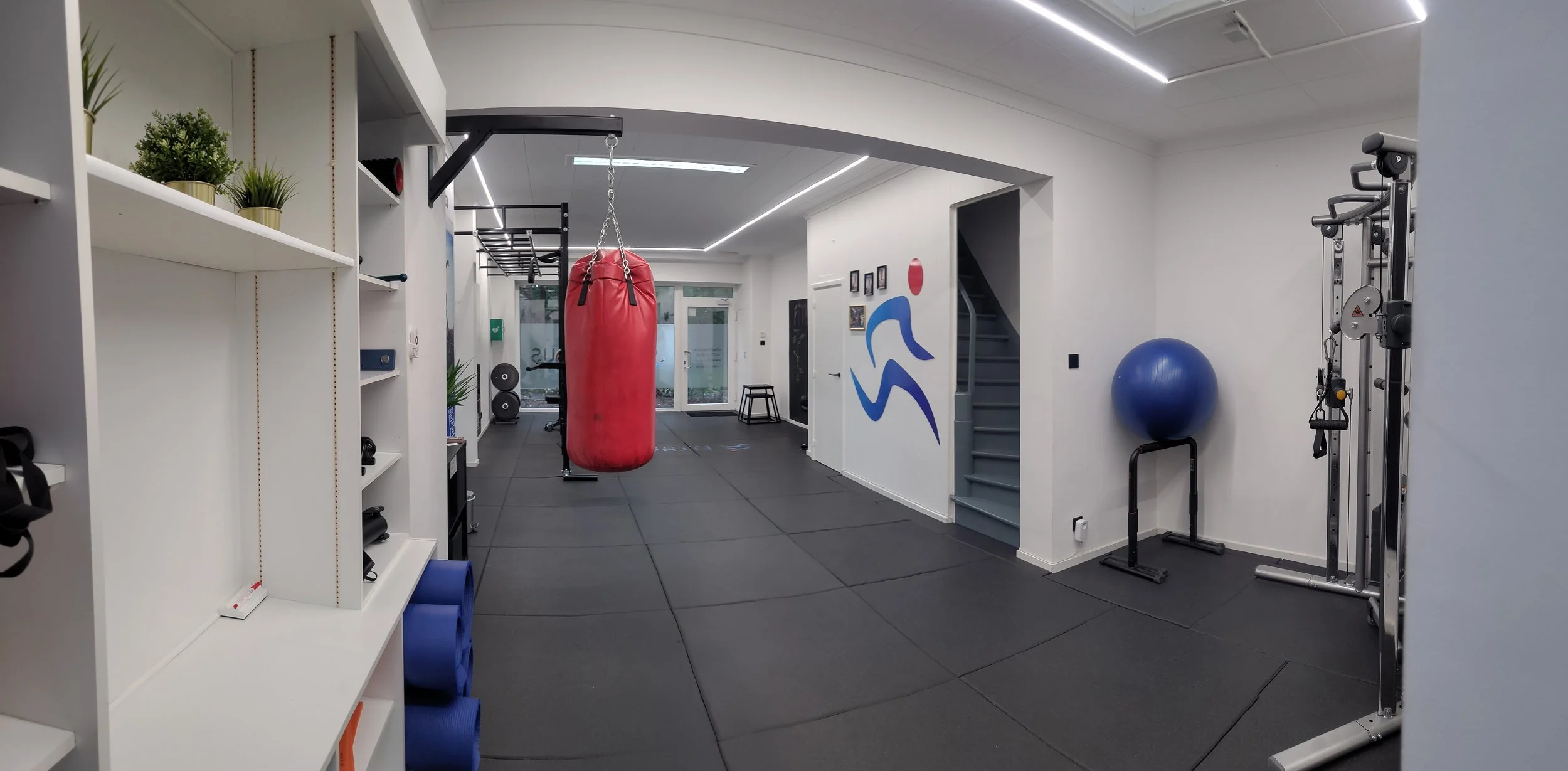 Binnen sportschool met punching bag, gewichten, fitnessapparaten, en trainingsmaterialen, wit muren en gelamineerde vloer.