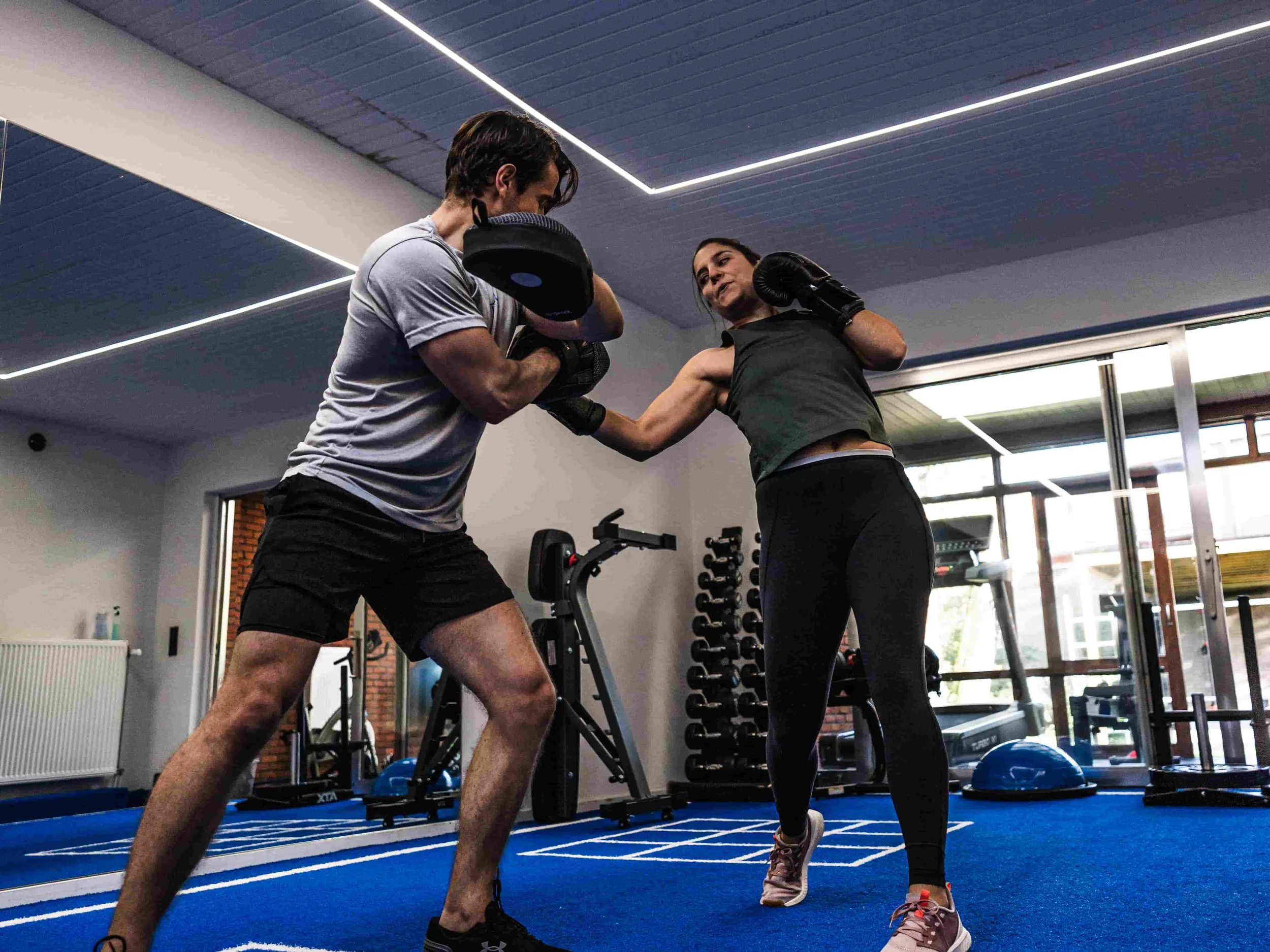 Twee mensen oefenen boksen in een sportschool, een man en een vrouw, terwijl de man in een bokshouding staat en de vrouw hem een stoot geeft.