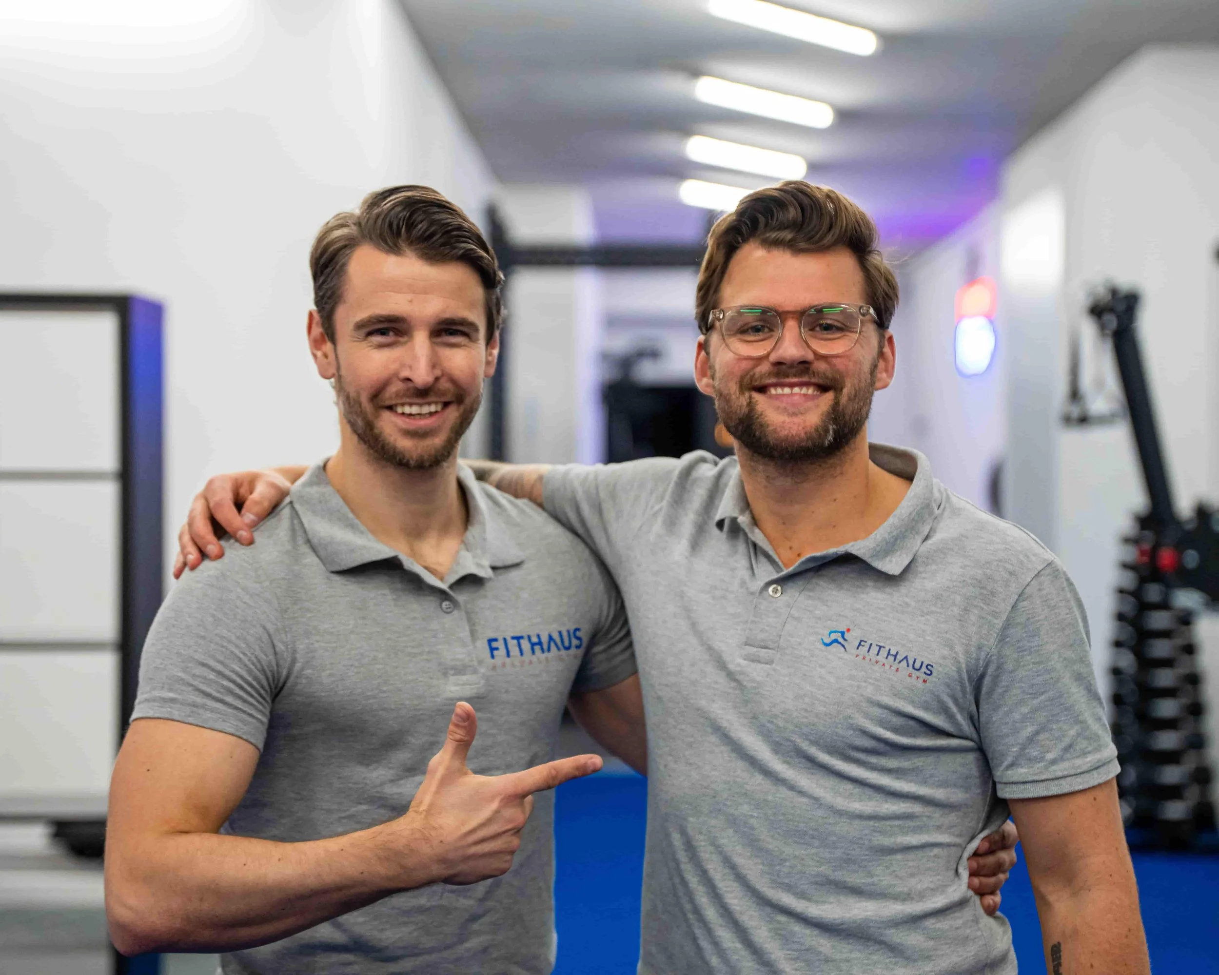Twee mannen in grijze shirts met het logo 'FITHAUS' staan samen in een sportschool, lachen en maken een handgebaar. Ze lijken vrienden of collega's.