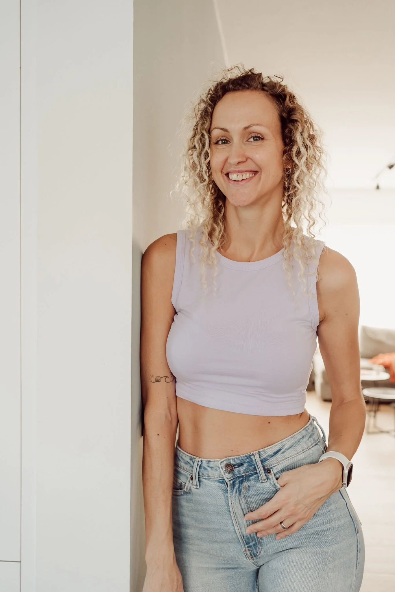 Een lachende vrouw met gekruld blond haar draagt een witte sporttop en spijkerbroek, leunt tegen een witte muur in een interieur met rustige kleuren.