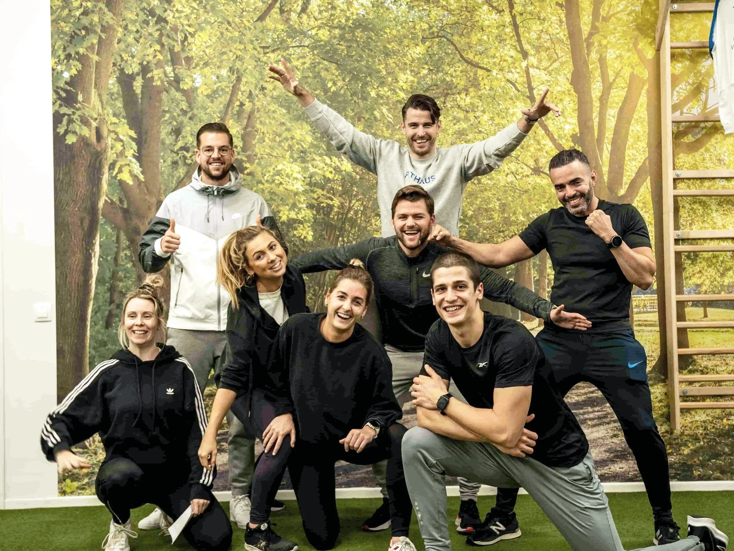 Groep enthousiaste mensen poseert in een bos, with enkele mensen knielen en anderen staan, om de groep te laten zien, sommigen maken gebaren en lachen, er is speelsheid en energie.