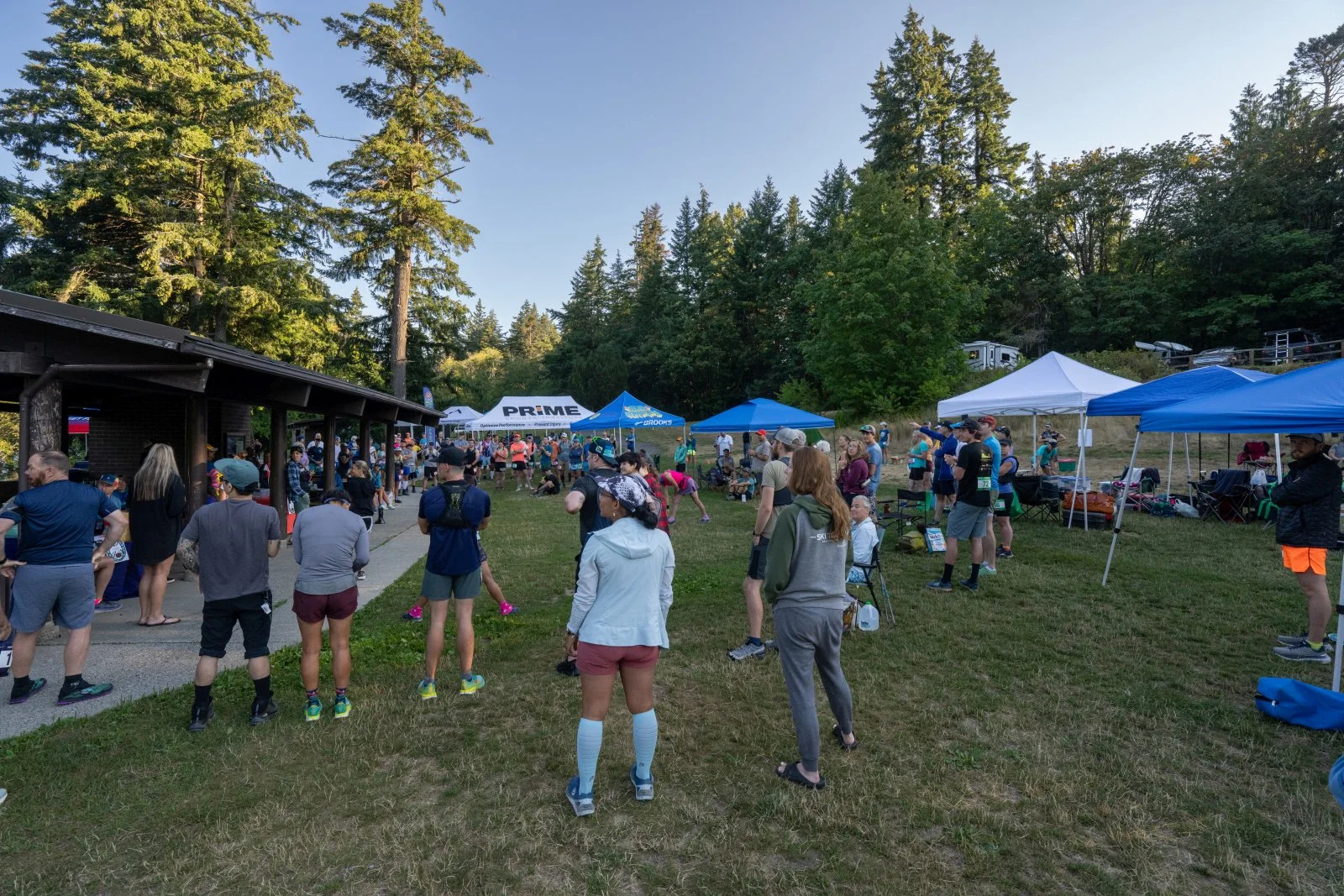 People gathered outdoors at a trail running event with tents, trees, and clear sky in the background.