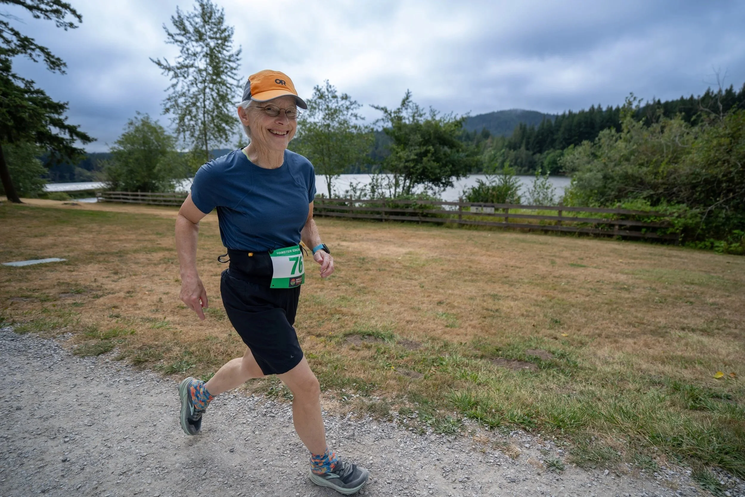 An elderly woman jogging outdoors by a lake, with mountains and trees in the background, wearing a blue shirt, running shorts, colorful socks, and a cap.