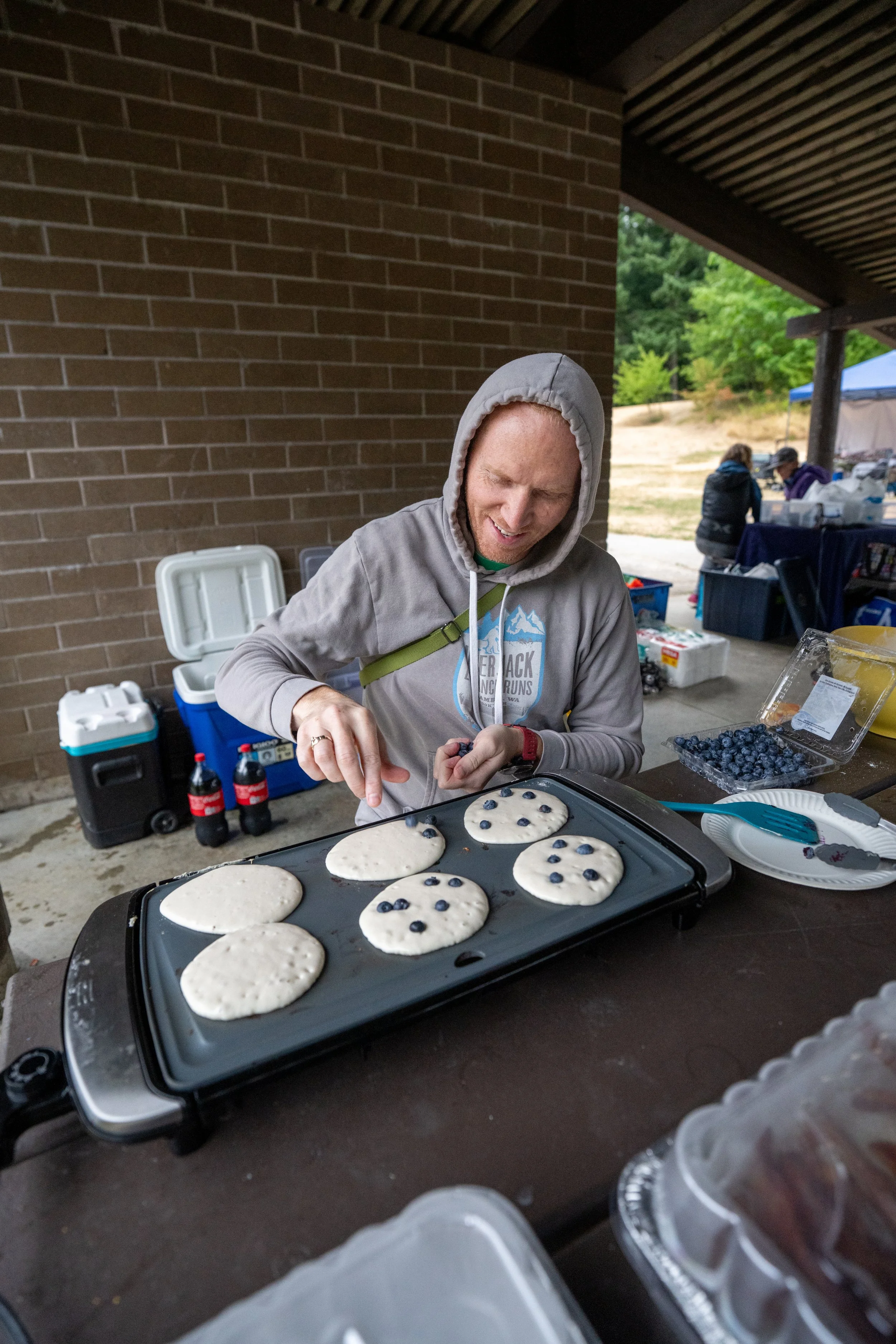 Person in hoodie making blueberry pancakes on electric griddle at outdoor event under a pavilion.