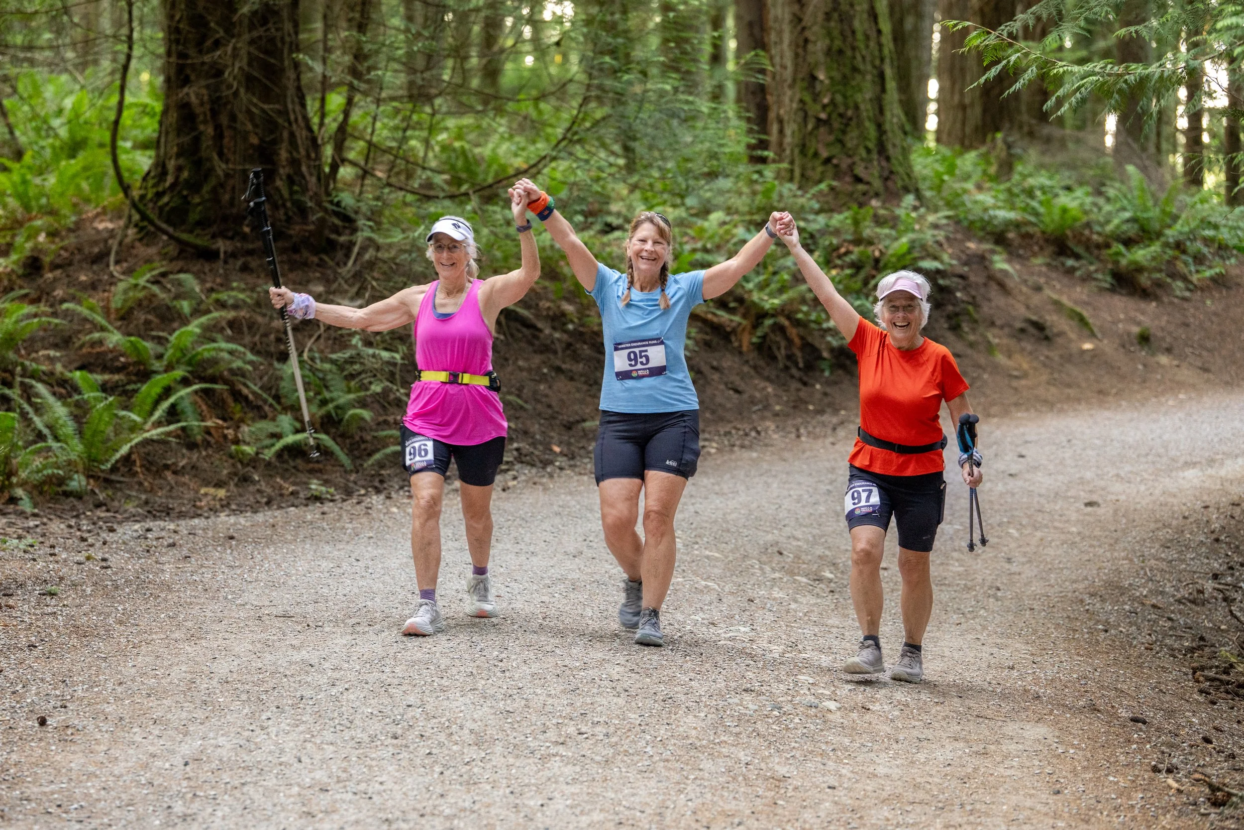 Three older women walking and holding hands on a forest trail, smiling and enjoying a day outdoors.