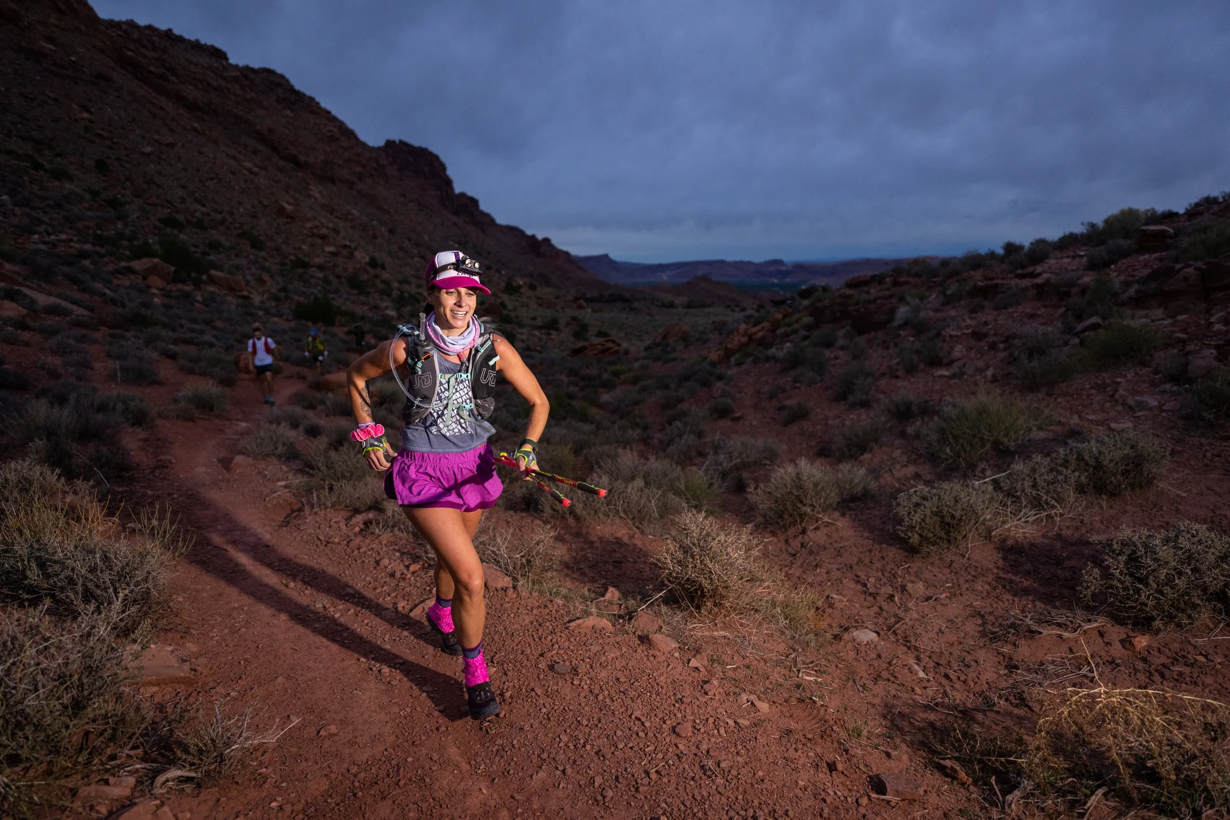 A woman trail running in a desert landscape at dusk, wearing pink shorts, a purple shirt, and a race bib, with a group of runners in the background and mountains under a cloudy sky.