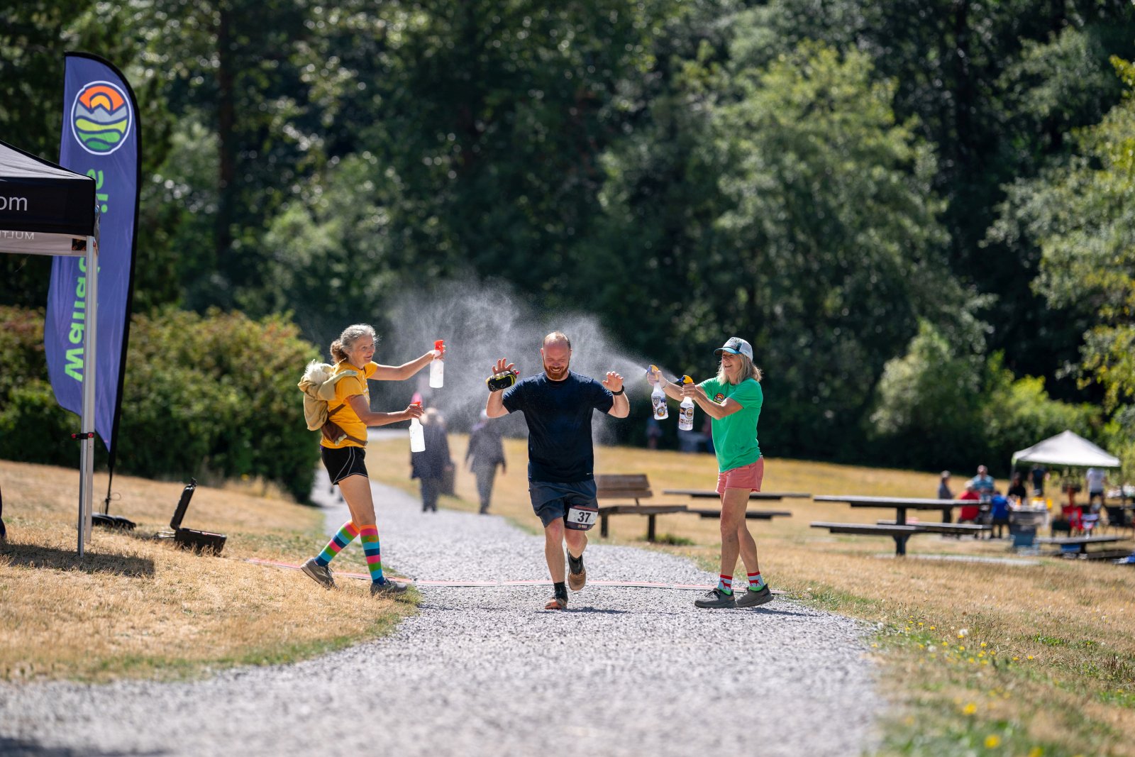 People celebrating and spraying water on a man near a finish line at an outdoor event on a sunny day, with trees and tents in the background.
