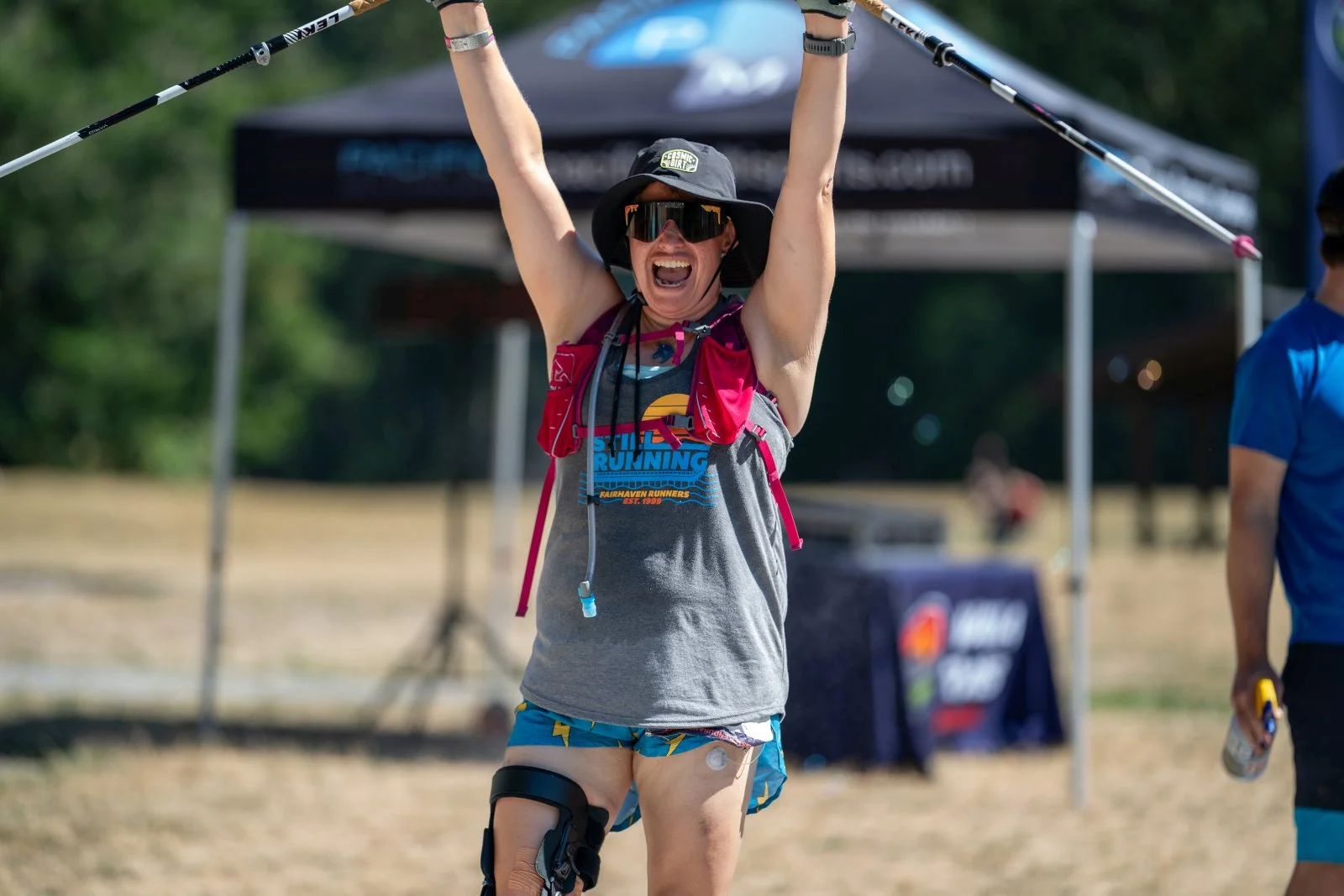 A woman wearing running gear, sunglasses, and a hat celebrates at an outdoor race event, raising her arms in victory.