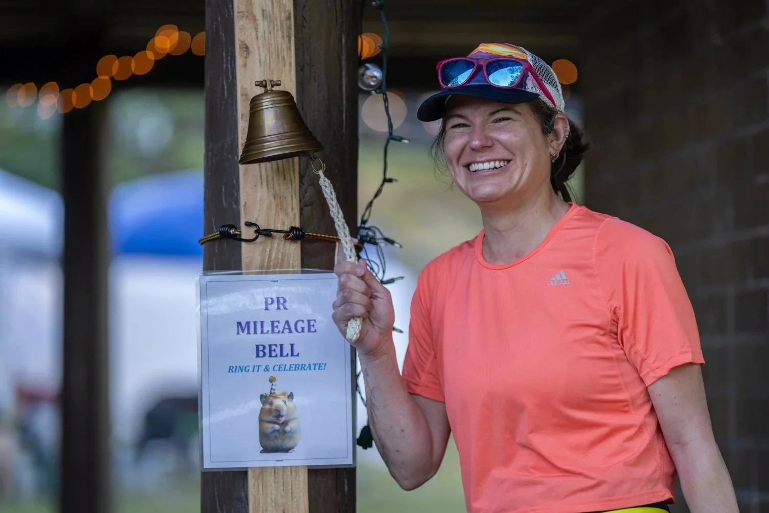 A woman in an orange athletic shirt and a baseball cap standing next to a sign that says 'PR Mileage Bell Ring it & Celebrate' with a picture of a hamster, holding a rope attached to a small bell, smiling.
