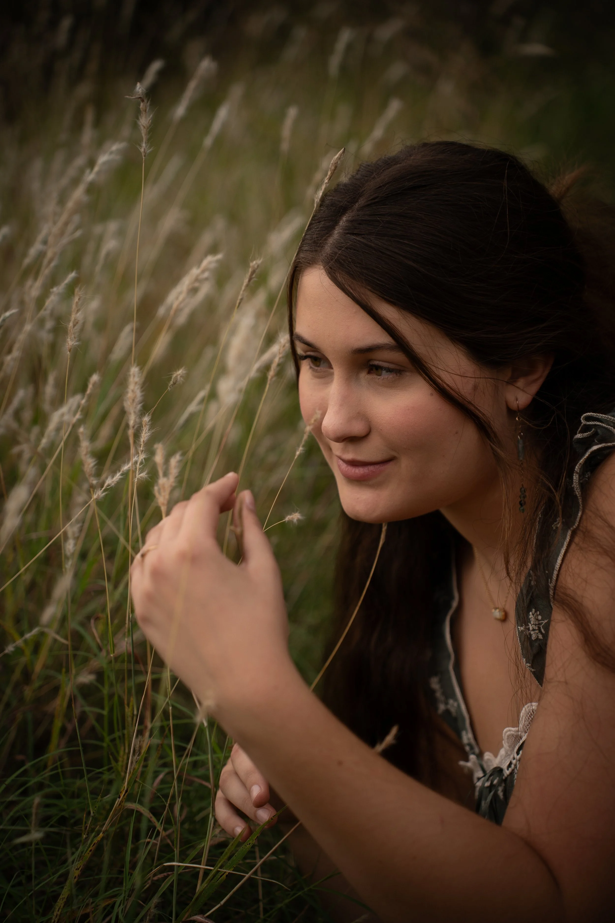 A young woman with long dark hair and light skin leaning in and smelling tall grass in a field, with a soft smile on her face.