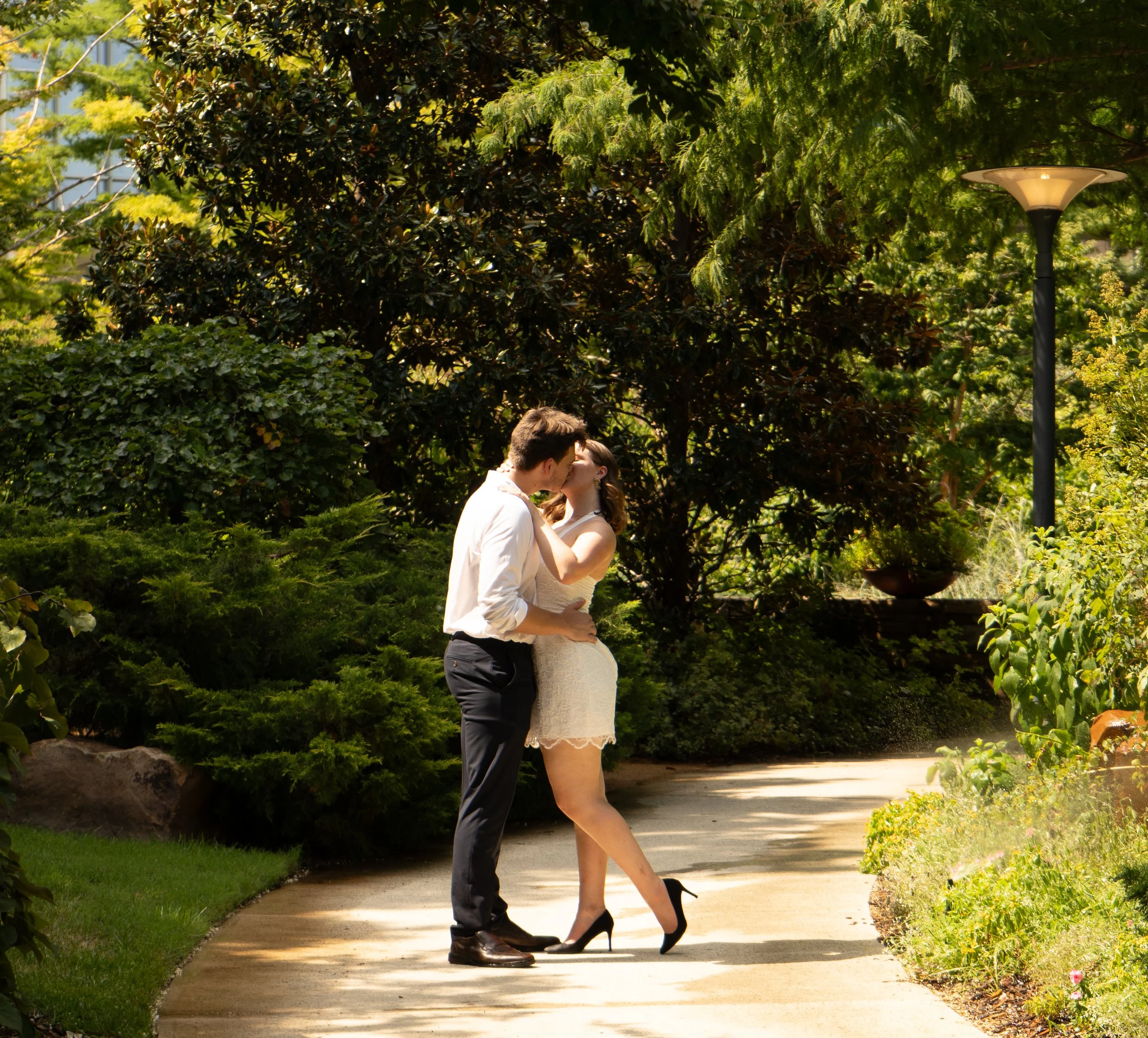 A couple kissing outdoors in a park, with lush green trees and shrubs around them, and a lamppost in the background.