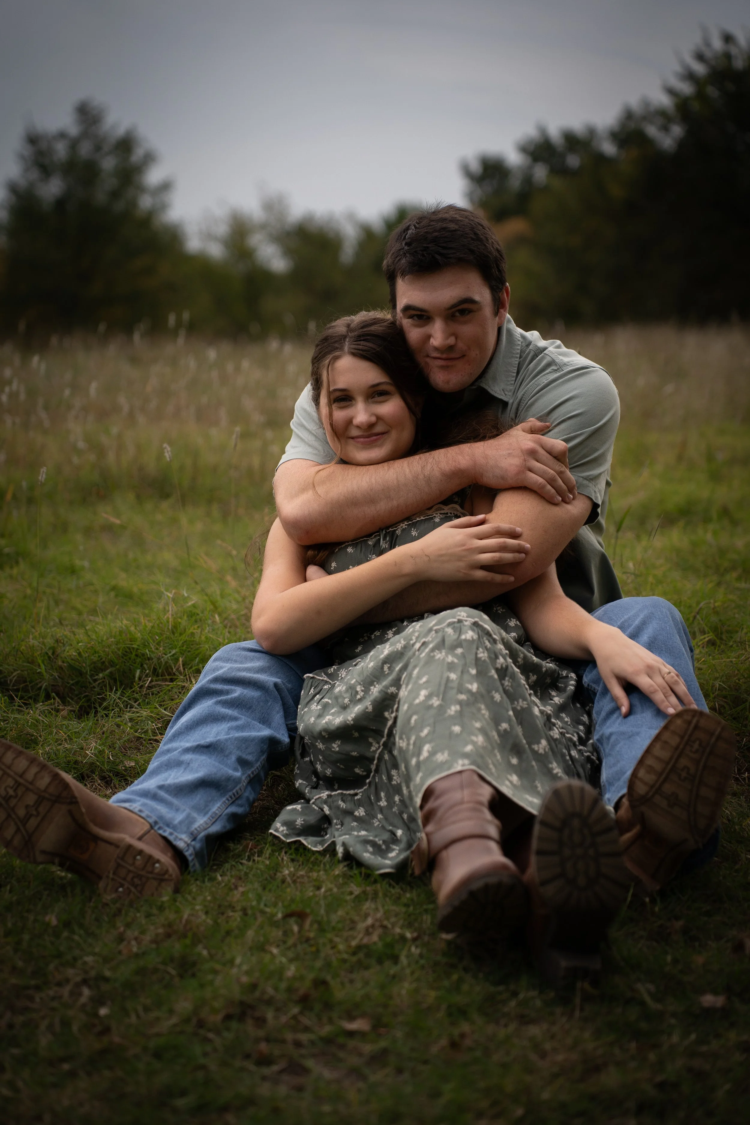 A young man and woman sitting on the grass in a field, hugging and smiling. Looks like they are outdoors during late afternoon or early evening.