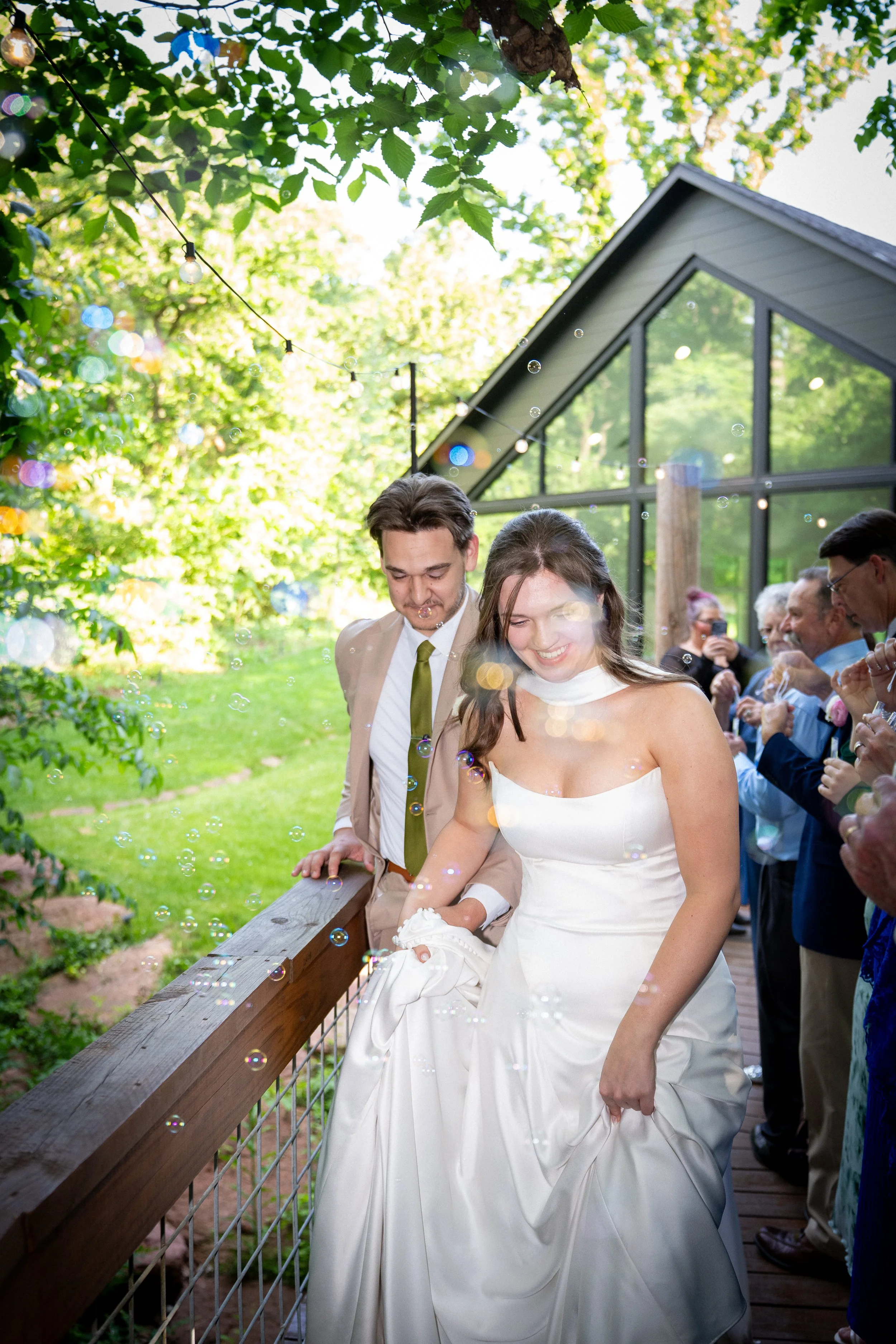 A wedding ceremony outdoors with a bride and groom standing on a wooden platform, surrounded by guests, while mist and soap bubbles float in the air. Green trees and a modern house with large windows are in the background.