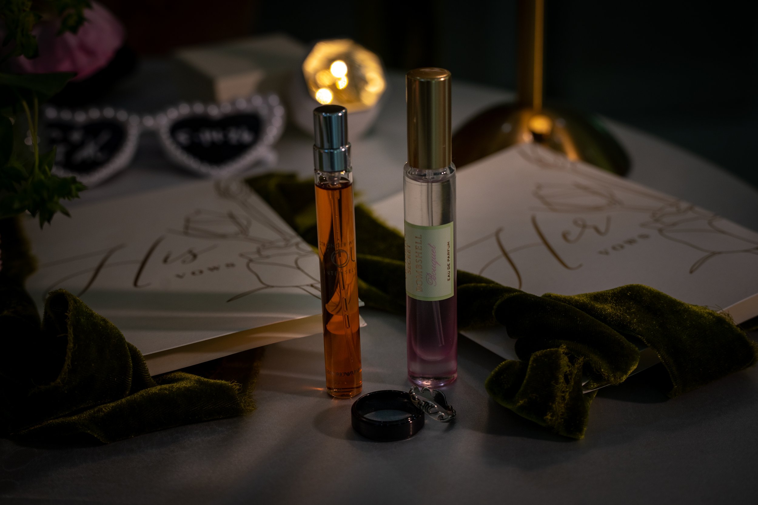 Two perfume bottles, one with orange liquid and the other with clear liquid, are placed on a table with a green velvet cloth. In the background, a pair of white-framed sunglasses with black lenses, a small round gold lamp, and some flowers are visibl
