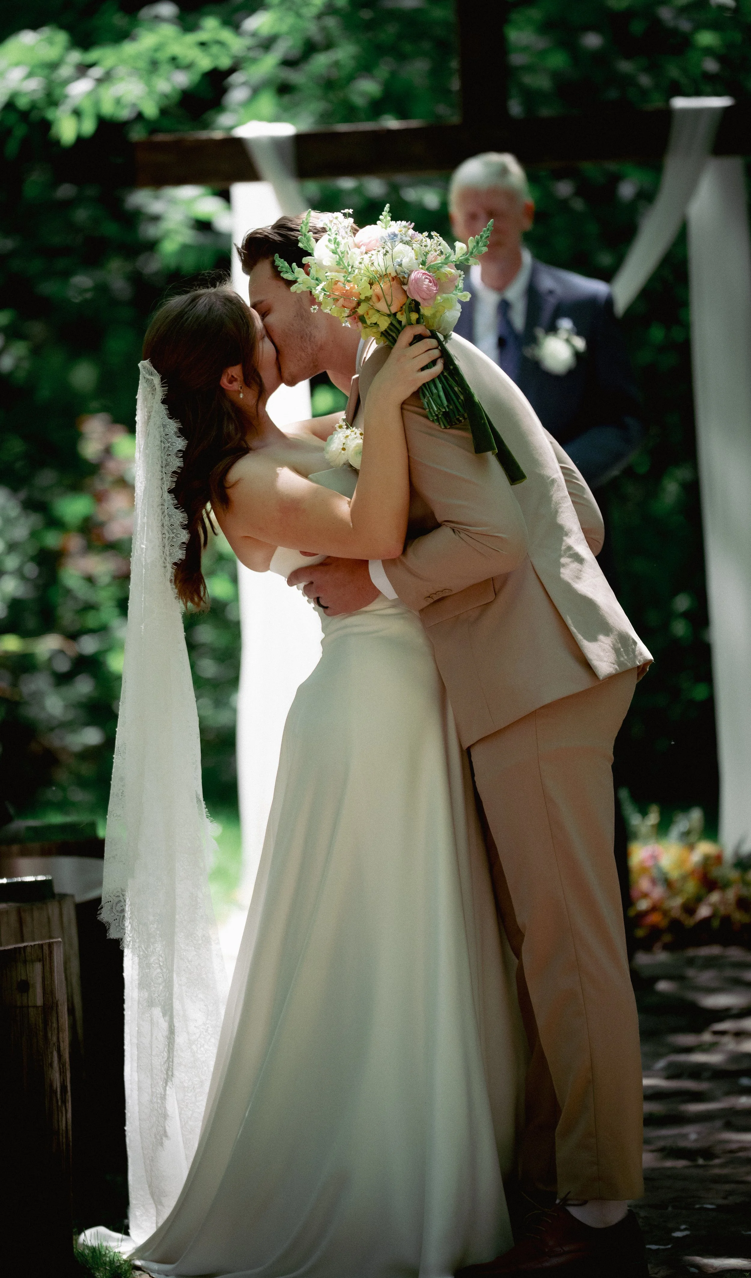 A bride and groom kiss during their outdoor wedding ceremony, with the bride holding a bouquet of pink and white flowers. The background has green foliage and a blurred officiant in a suit watching them.