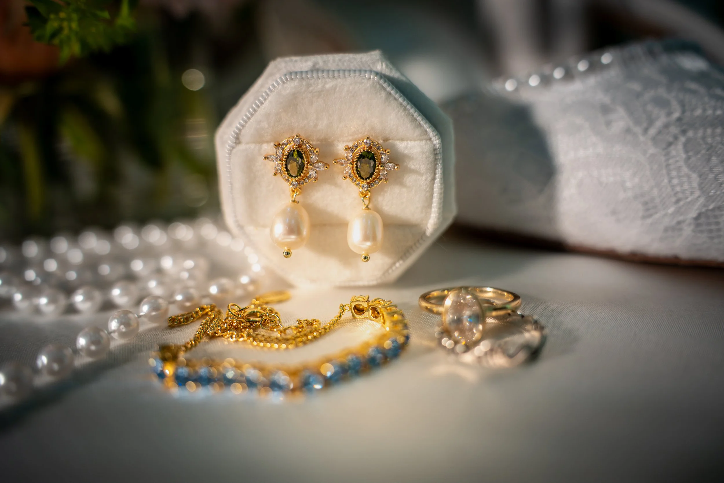 Close-up of gold earrings with dark green gemstones and pearl drops inside an open white jewelry box, surrounded by gold jewelry, pearls, and a gray and white textured fabric.