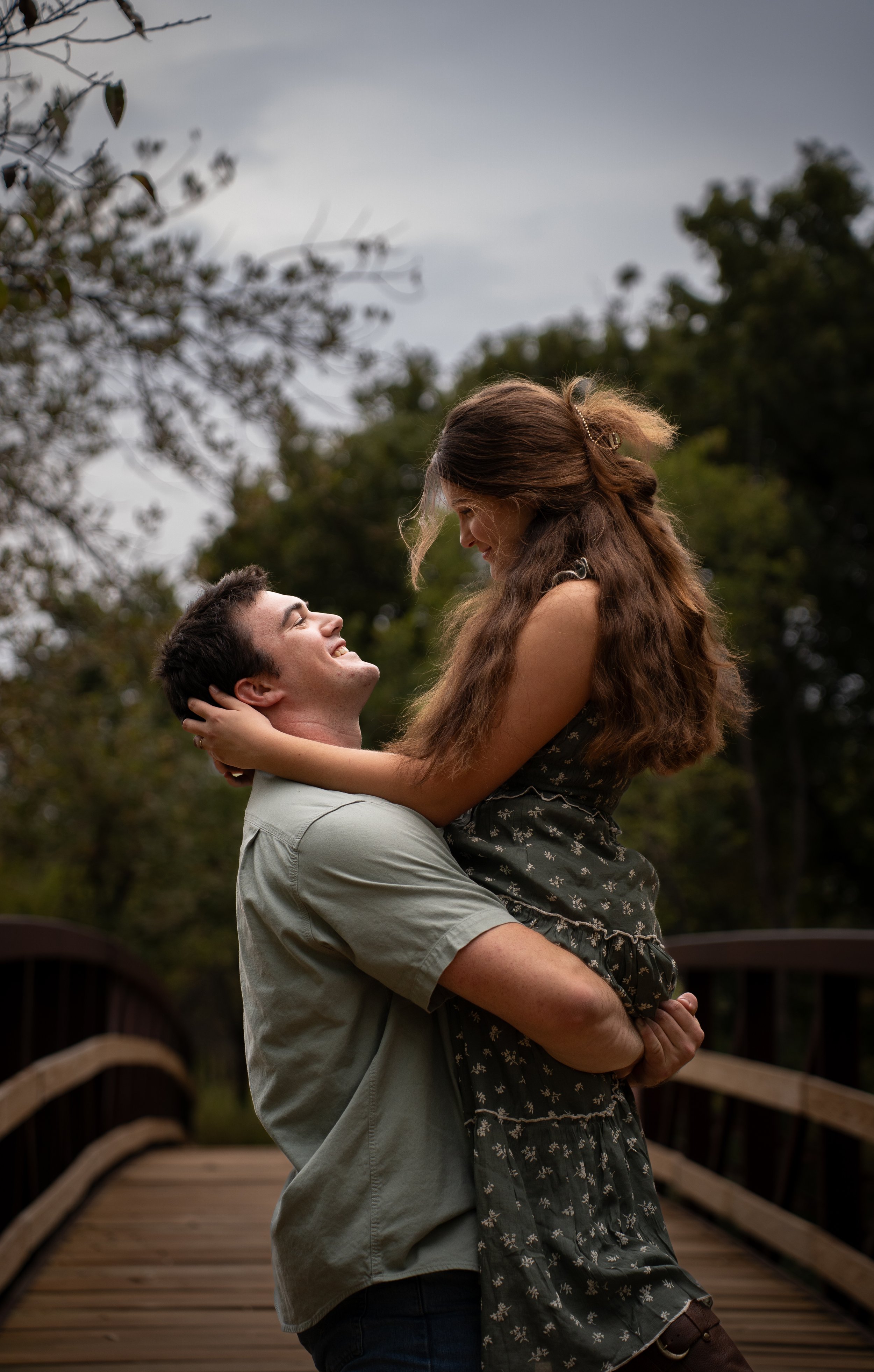 A man lifting a woman in an outdoor setting with trees in the background, both smiling and looking at each other.