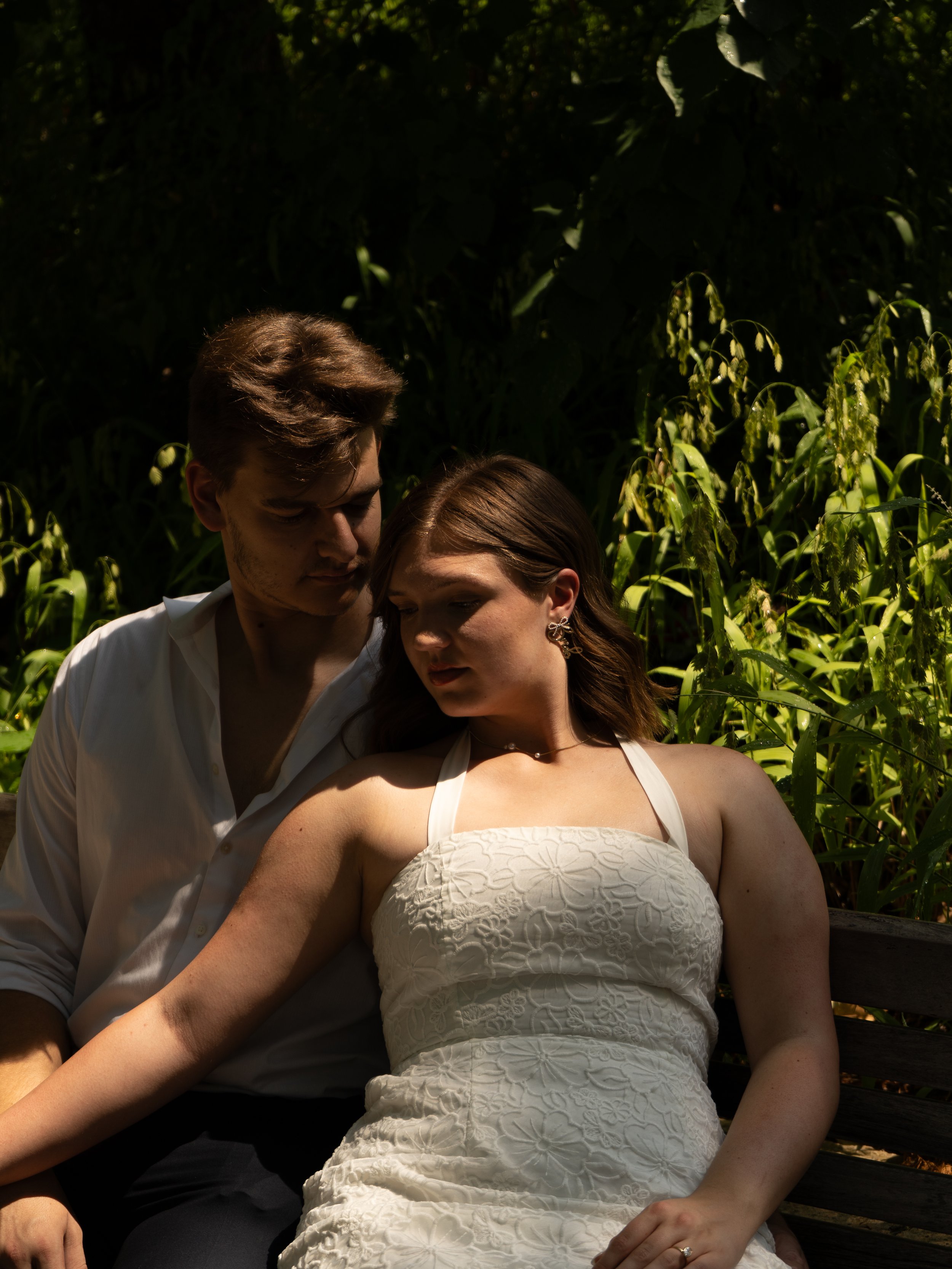 A young couple sitting close together outdoors on a wooden bench, surrounded by lush green foliage and trees, with sunlight filtering through.