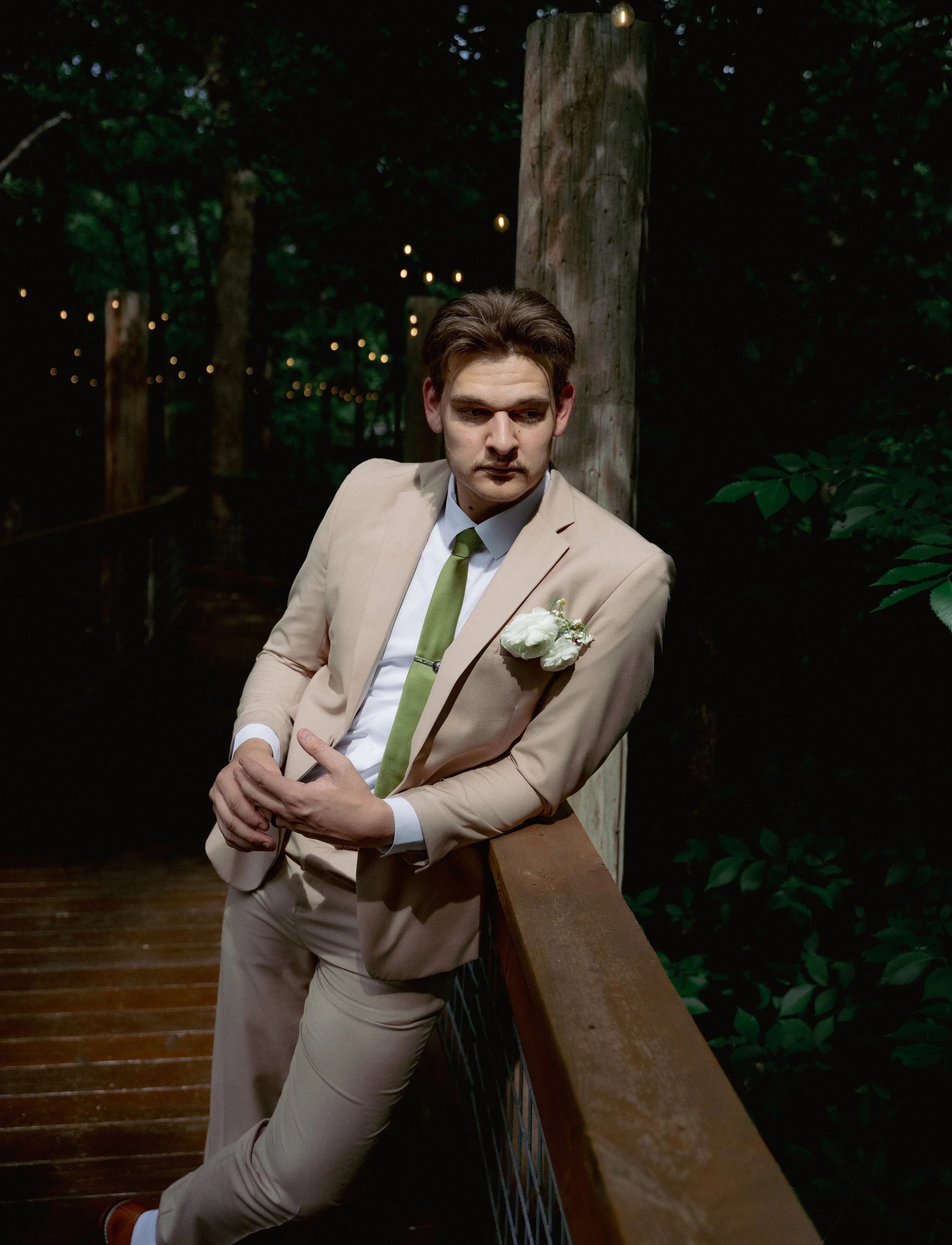 A young man in a beige suit with a light green tie and white shirt posing outdoors at night on a wooden bridge with a blurred background of trees and string lights, leaning against the railing with one arm resting on it.
